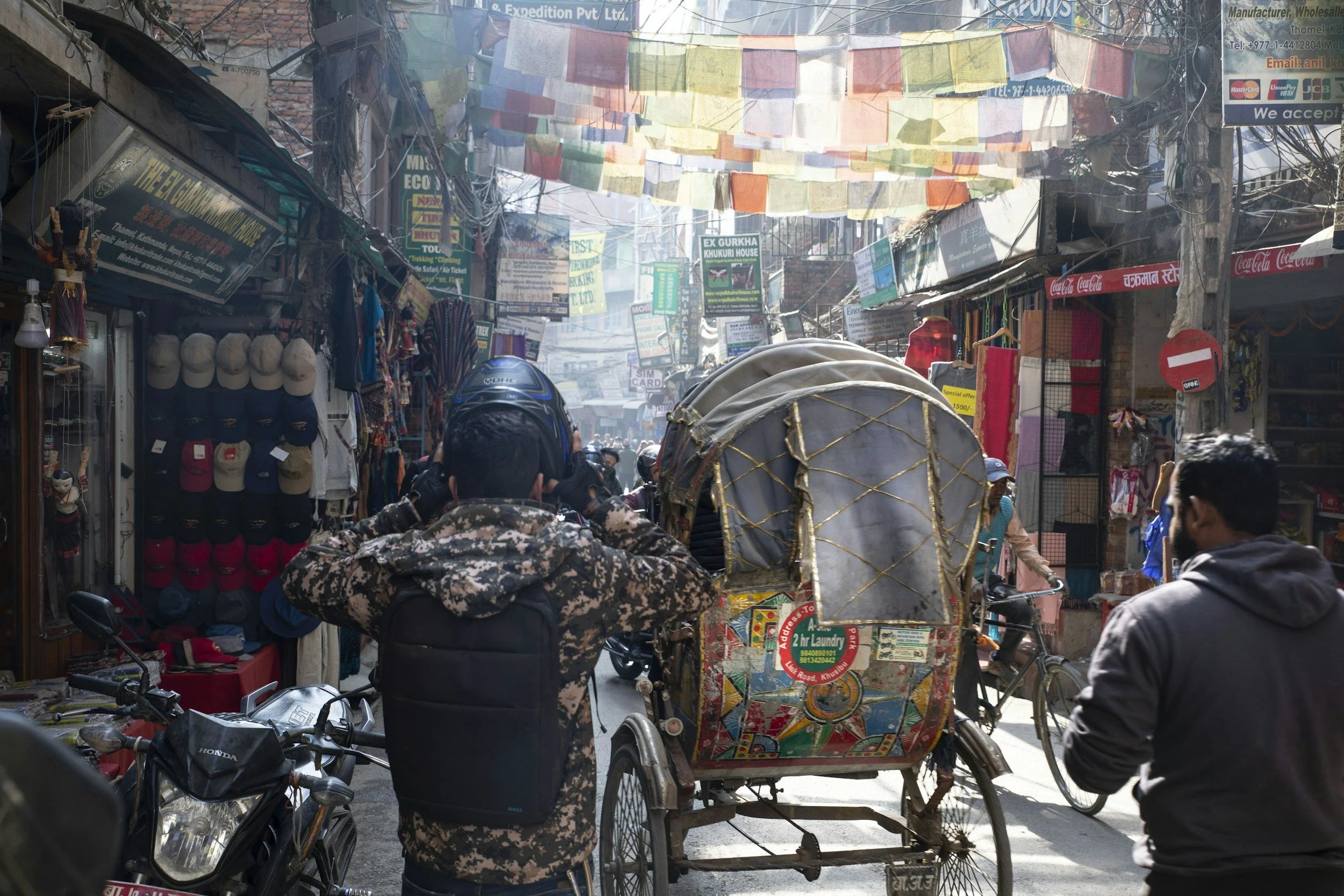 A rickshaw moving through the busy streets of Thamel, with prayer flags overhead and shops, locals, and tourists lining the street.