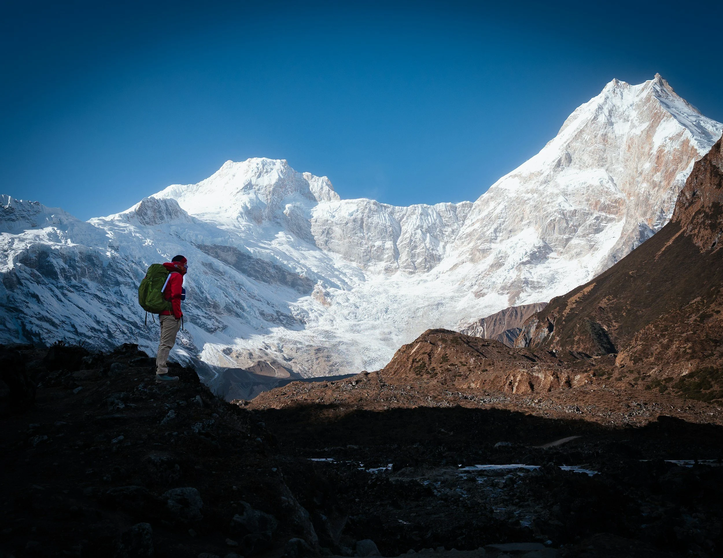 A trekker stands in front of the massive Manaslu peak, with the towering Himalayan mountain dominating the background.