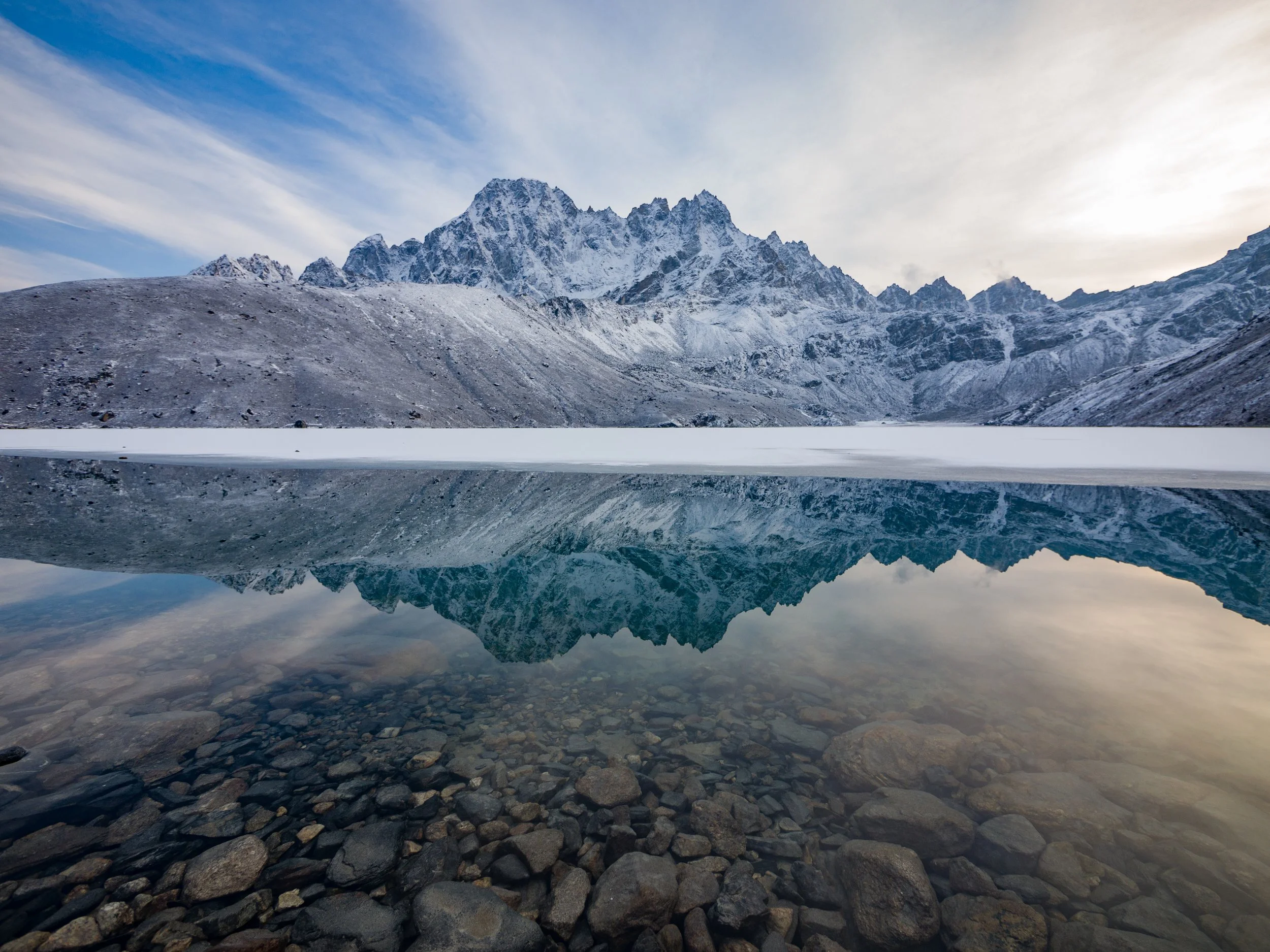 Snow-covered mountains reflected in a calm lake with rocky shoreline, blue sky, and thin clouds.
