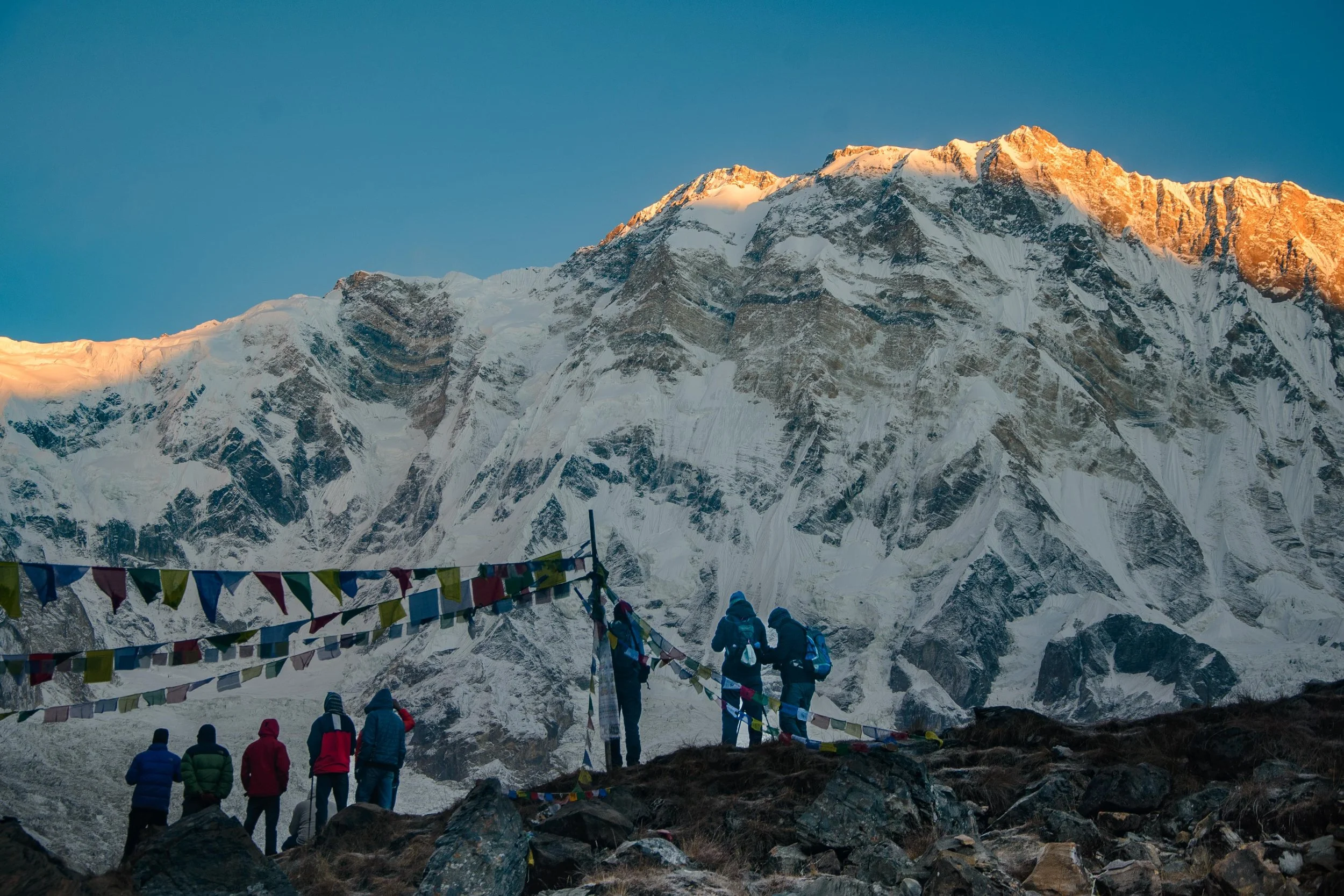 Trekkers waiting for the first sunlight to hit the mountain tops, with Annapurna I in the background and prayer flags and trekkers in the foreground.