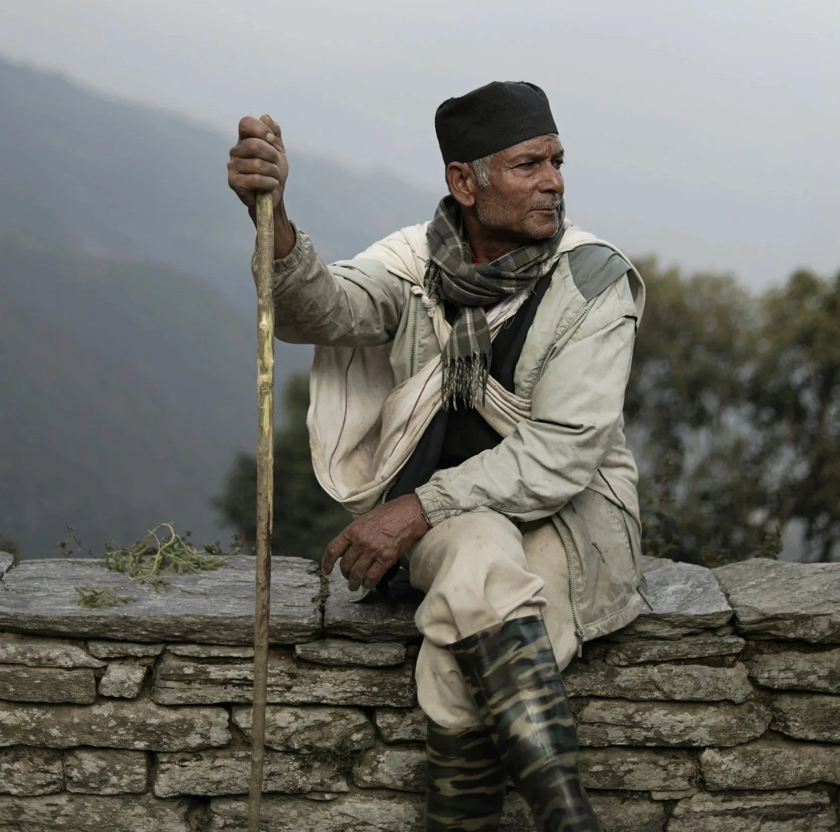 Local man resting on a stone wall with a wooden walking stick along a mountain trail in Nepal
