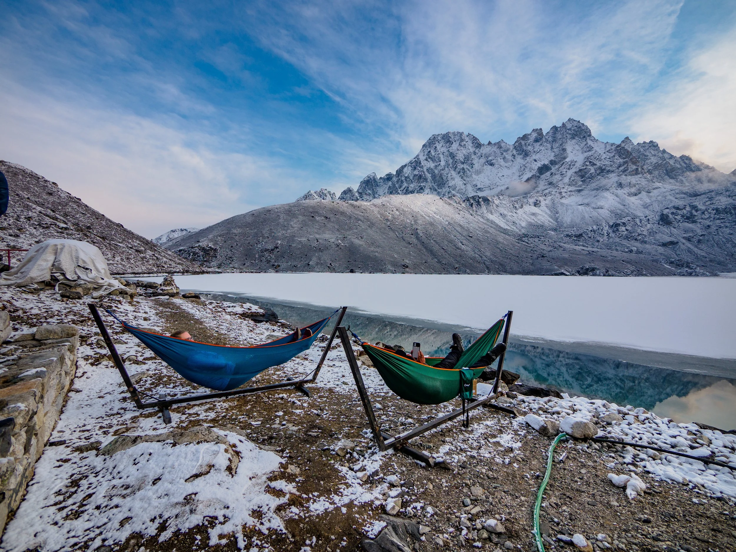 Two hammocks on snowy ground beside a partially frozen lake with mountains in the background.