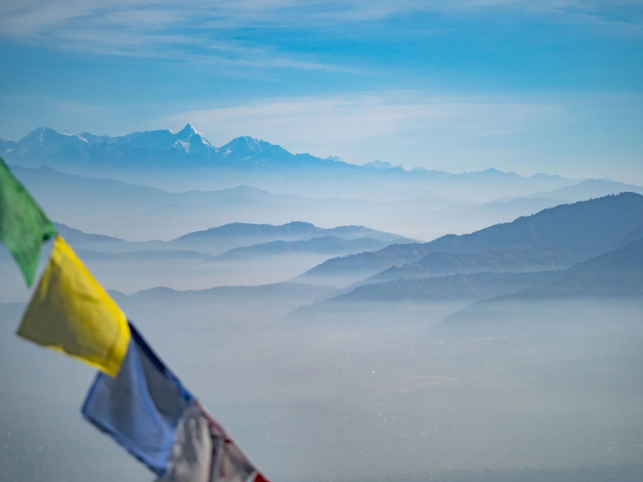 Mountain range shrouded in mist with colorful prayer flags in the foreground and a clear blue sky.