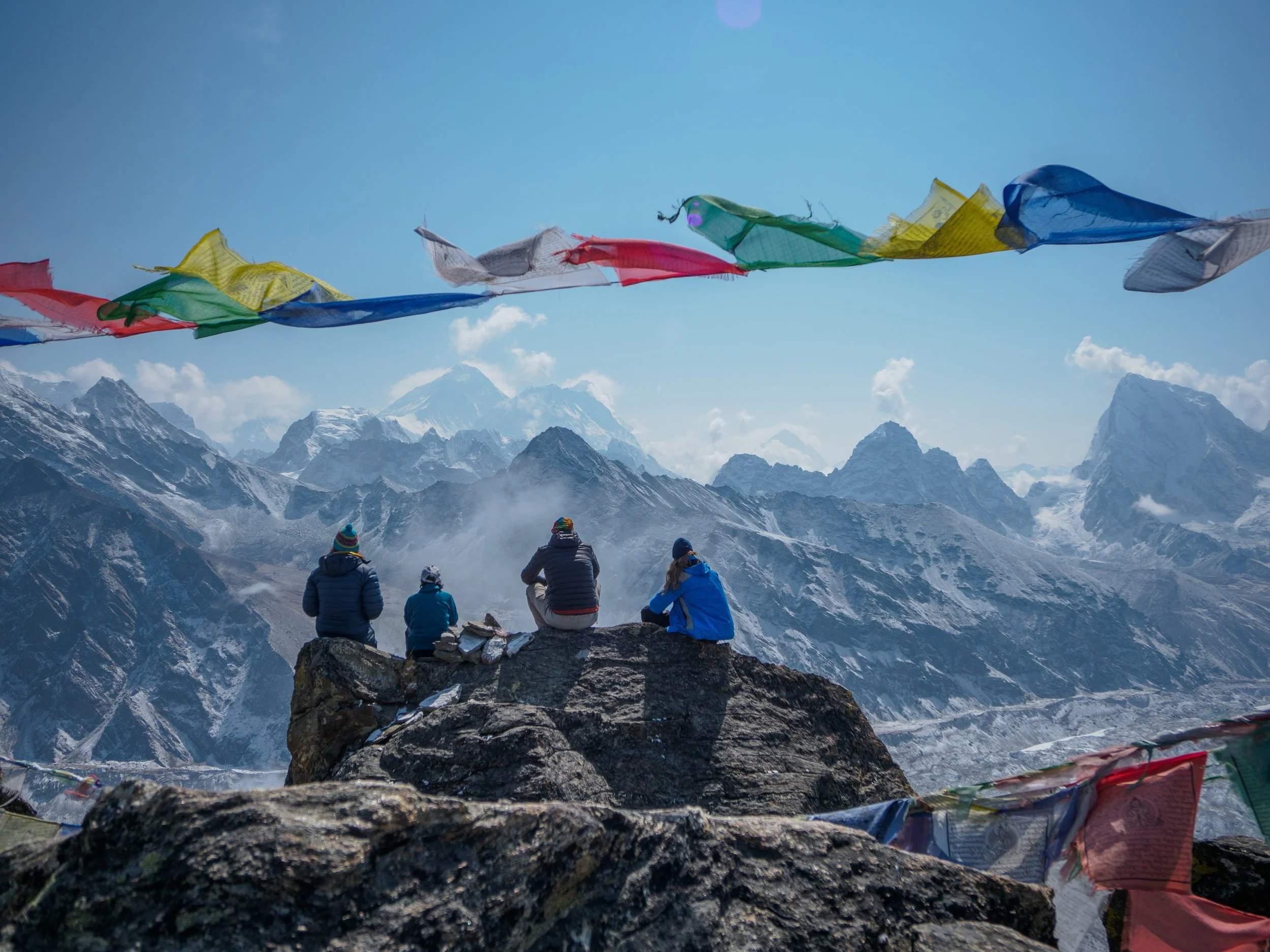 Four people sitting on a rocky outcrop overlooking snowy mountains with colorful prayer flags in the foreground.