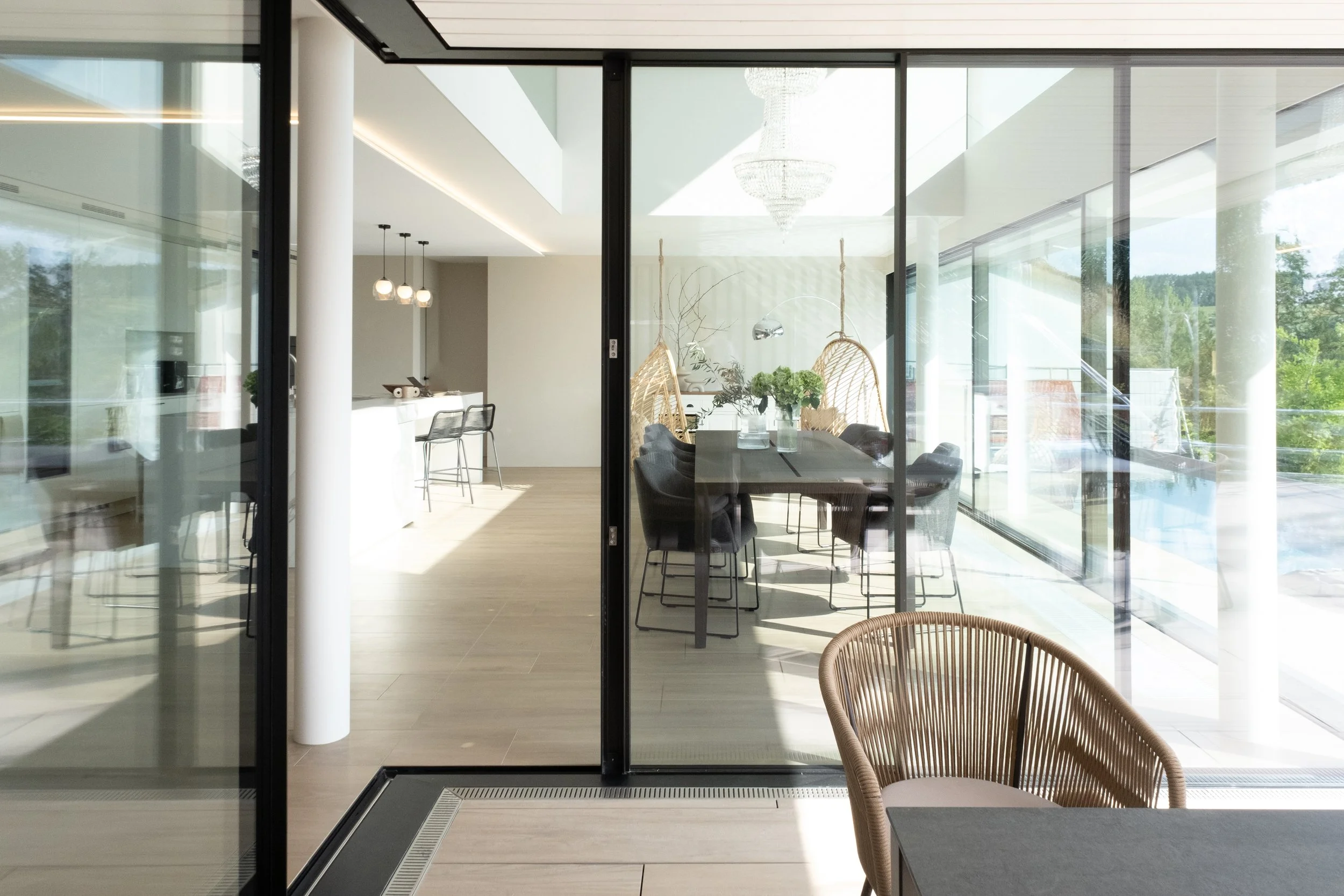 Modern dining area with black chairs and a long table, bright sunlight, glass walls, and a view of greenery outside.