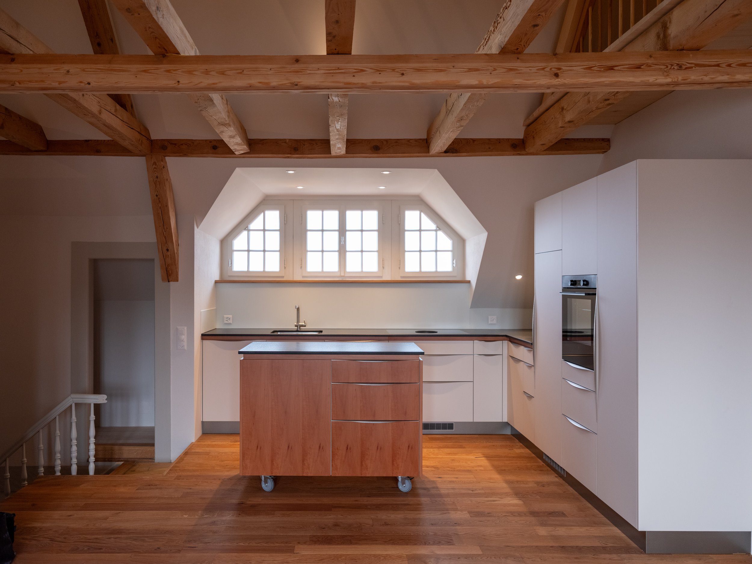 Modern kitchen with white cabinets, a wooden island on wheels, and a large window with multiple panes in an attic space with wooden ceiling beams and hardwood flooring.