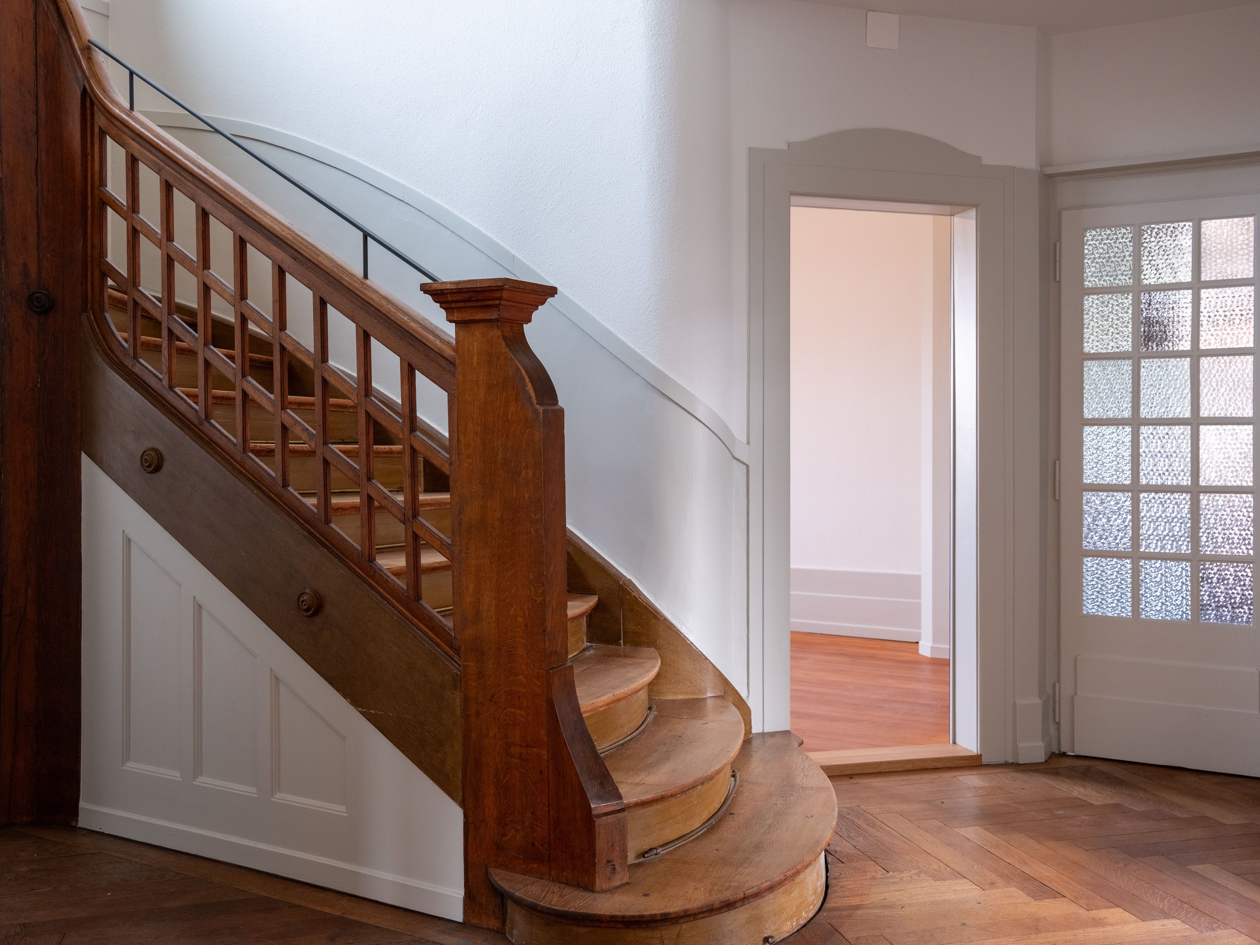 Interior view of a staircase with wooden handrail and steps, adjacent to a doorway with a glass-panel door, and hardwood flooring.