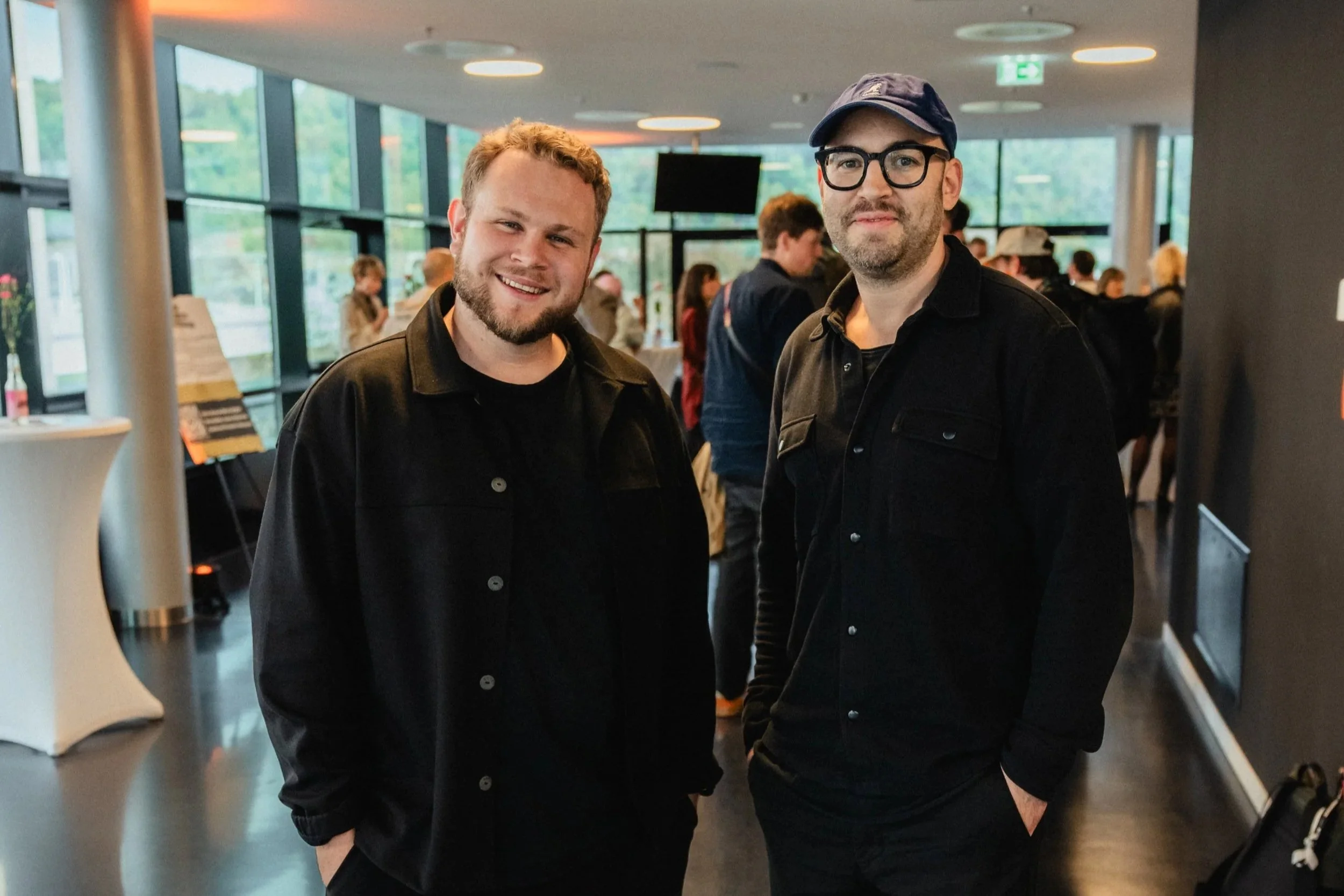 Zwei Männer im modernen Foyer bei einer Veranstaltung in Mainz, beide in schwarzer Kleidung lächelnd vor einer Gruppe von Gästen – Team der Eventagentur KULTUR & VERGNÜGEN.