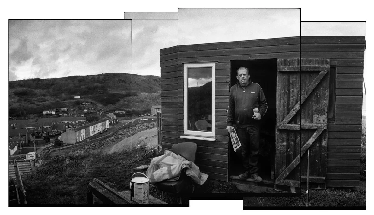Black and white photo of a man standing in the doorway of a small wooden house, holding a cup and newspaper, with a view of a hilly town in the background.
