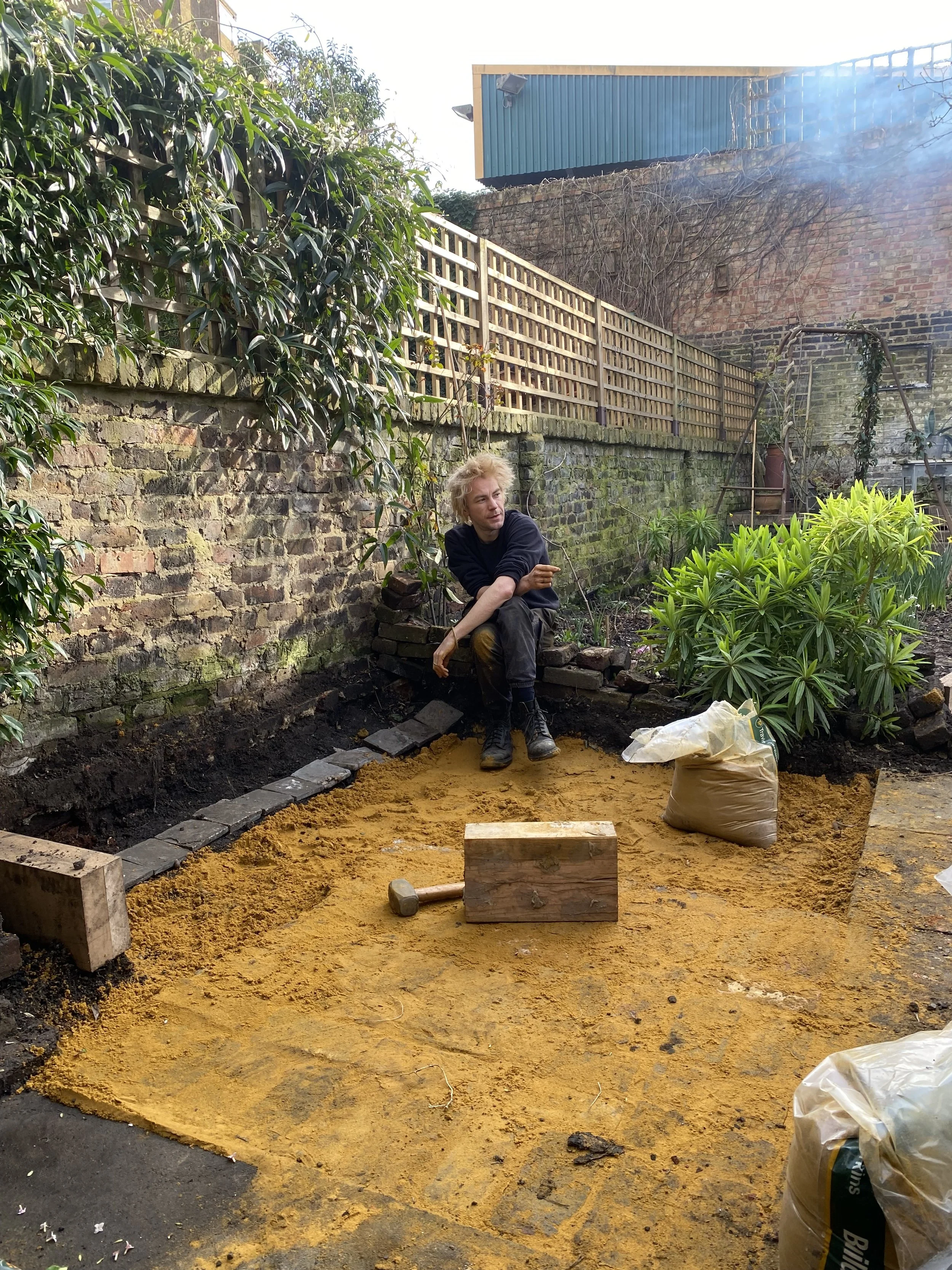A person with blond curly hair sitting on a small brick wall in a garden, surrounded by green bushes and dirt. They are holding a small gardening tool and appear to be working on the garden bed. There are bags of soil, a wooden block, and a hammer ne
