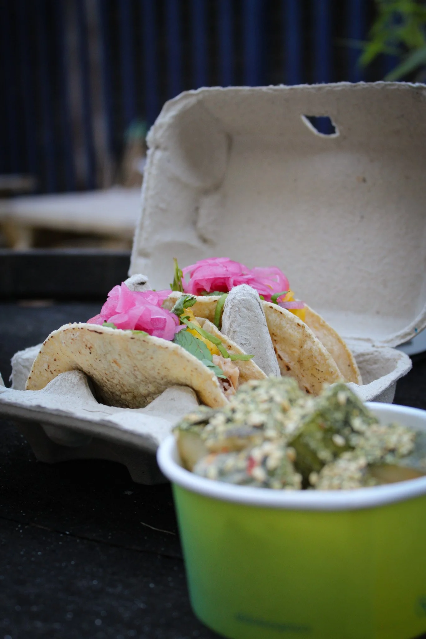 A takeout container with tacos and a cup of guacamole on a black surface.
