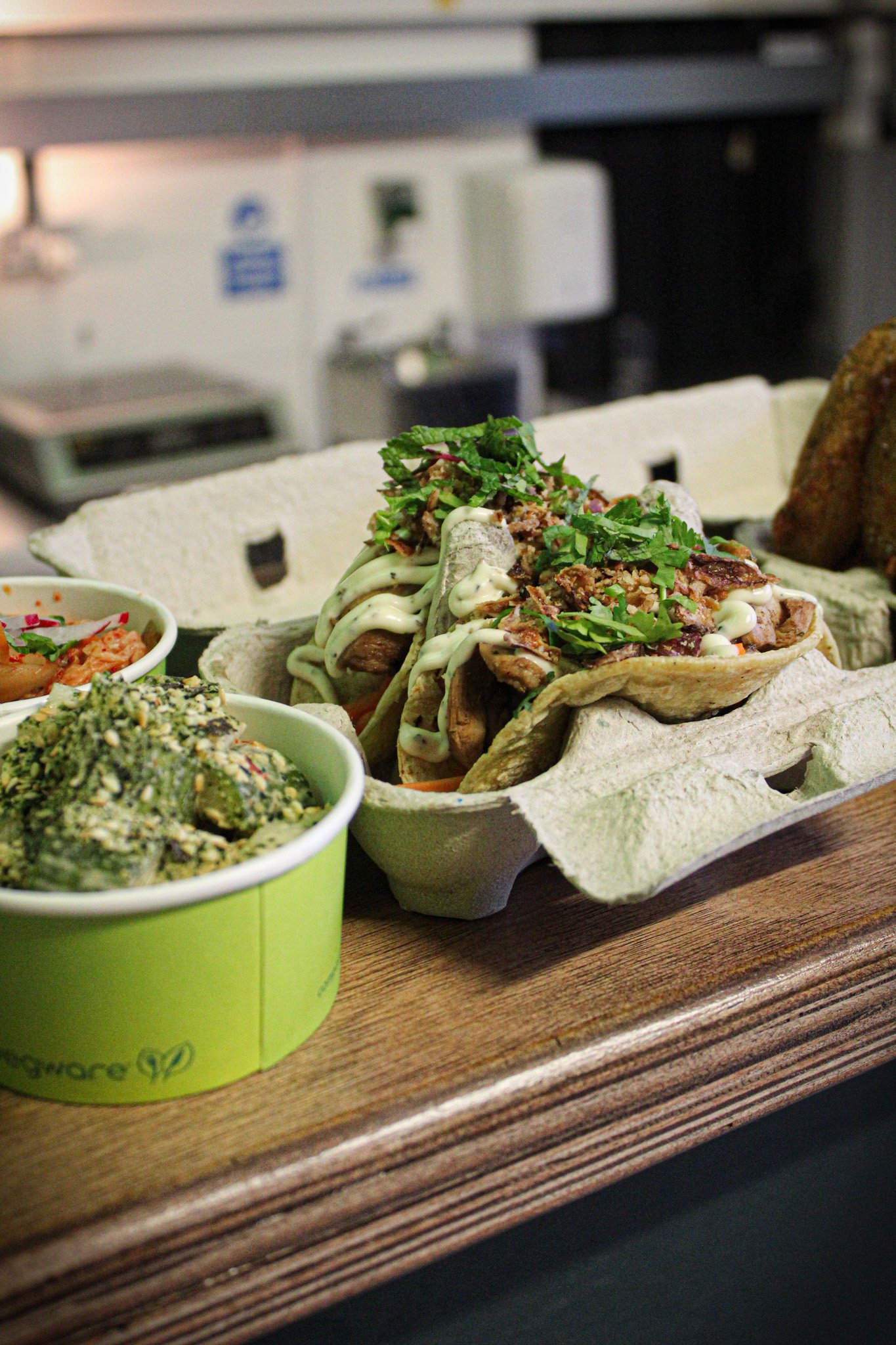 Two tacos filled with meat, cilantro, and sauce, served in a paper tray on a wooden surface, with a side of green vegetables and rice in green containers.