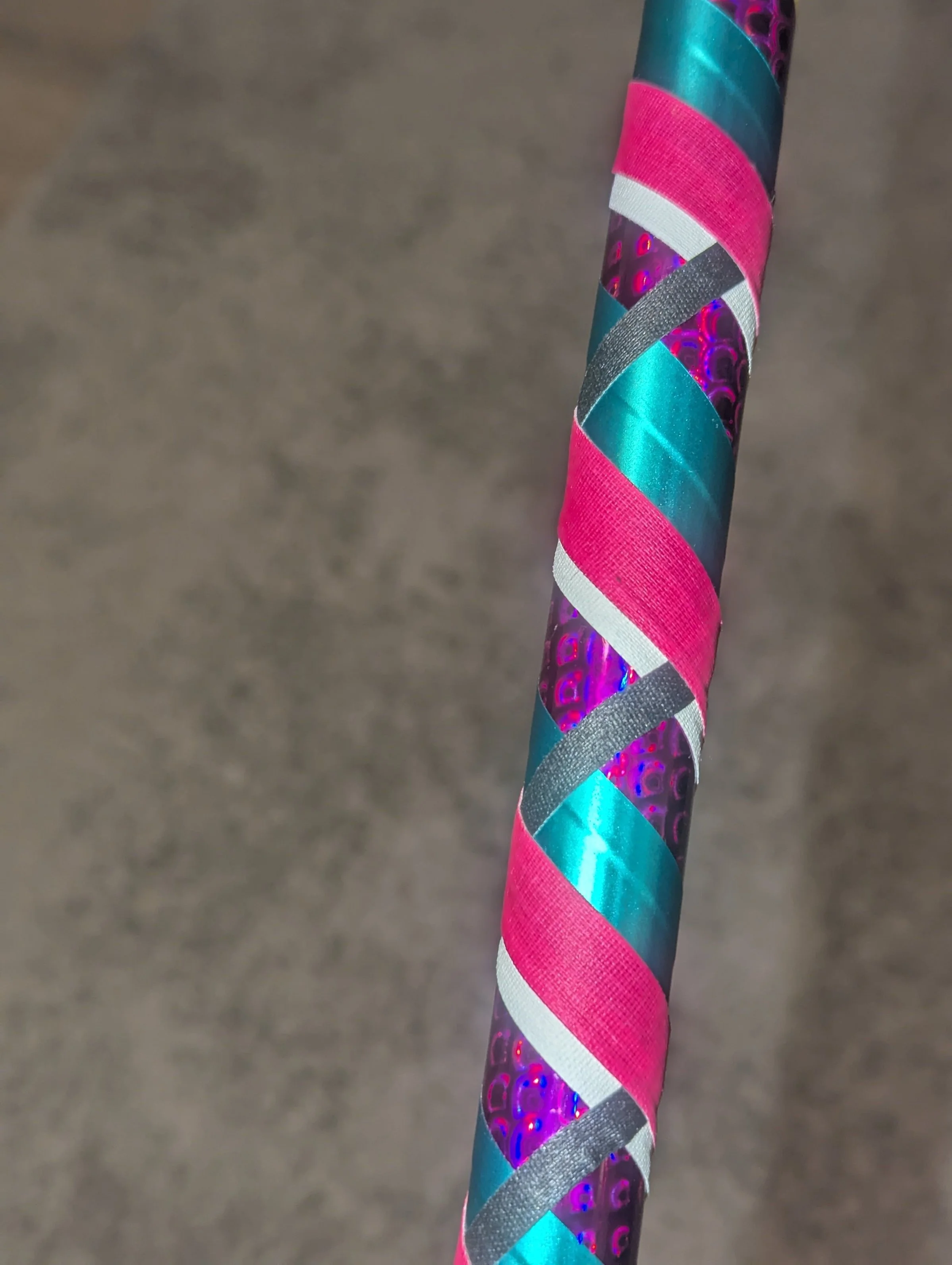 Nahaufnahme eines mit bunten Bändern umwickelten Schlauch oder Stocks. Die Bänder sind in metallisch glänzendem Blau, Pink und Rosa angeordnet, mit Mustern und texturierten Oberflächen.