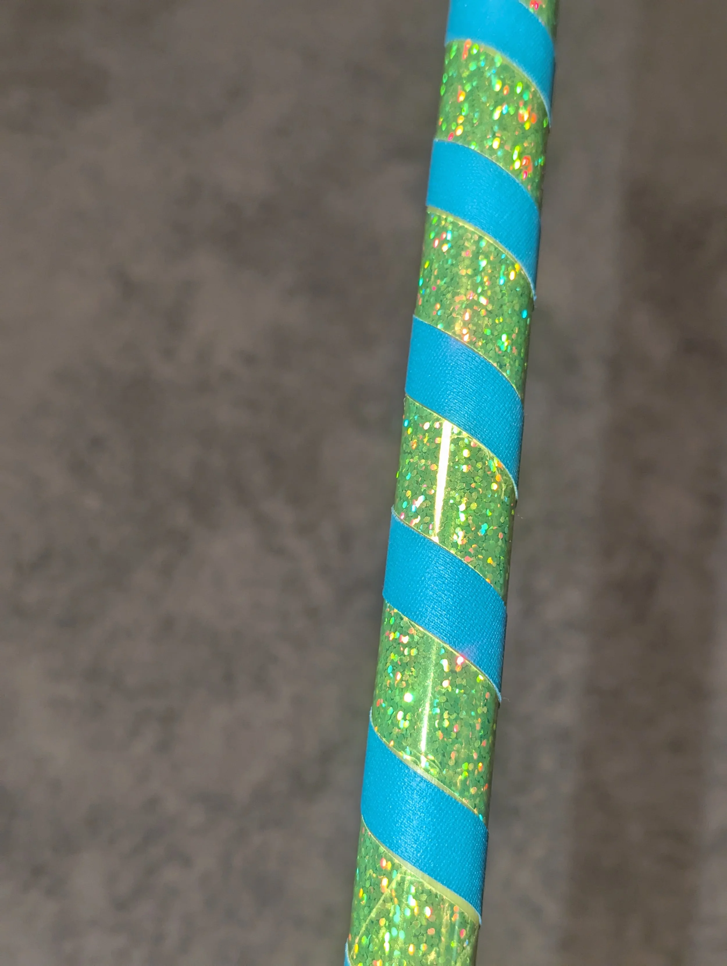 Ein grüner Glitzerstab mit spiralförmigem, blauem Band umwickelt auf grauer Fläche.