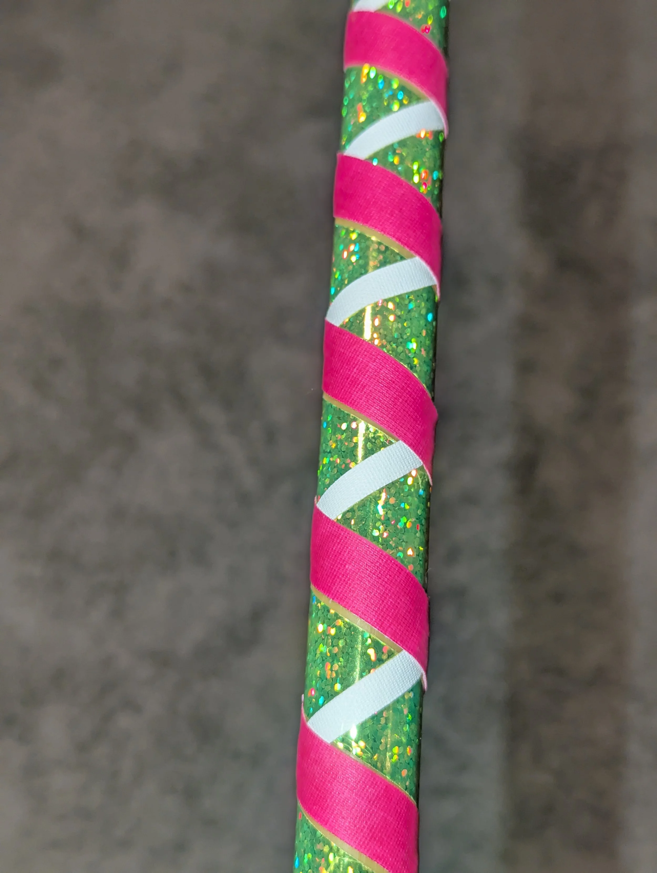 Nahaufnahme eines glitzernden grünen Stocks, der diagonal mit rosa und weißen Bändern umwickelt ist, vor einem grauen Hintergrund.