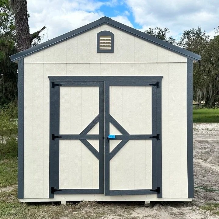 Small outdoor shed with beige walls, dark gray trim, double doors with diagonal support beams, and a small vent on the gable end.
