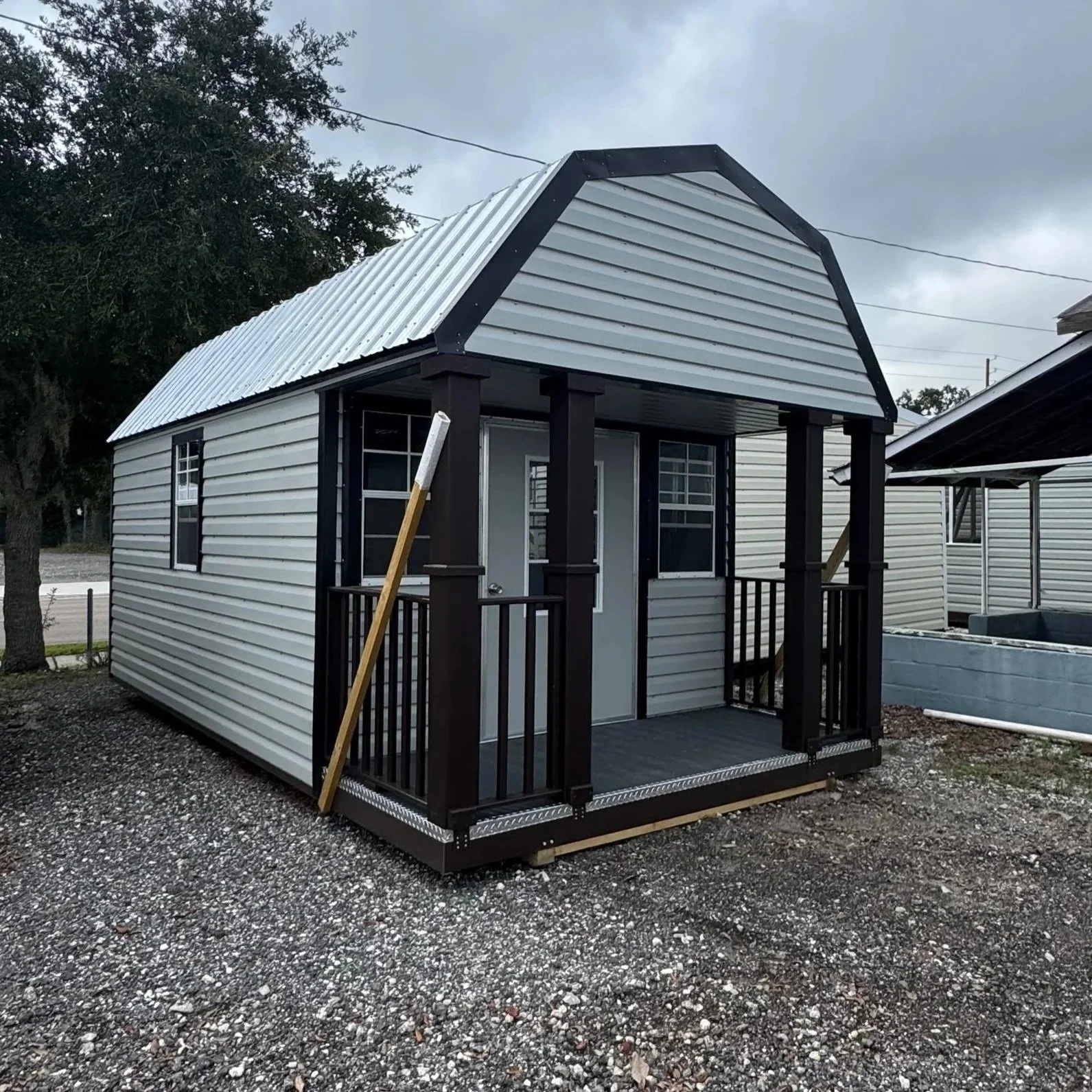 Small white metal shed with black trim and a small porch, on a gravel lot under cloudy sky.