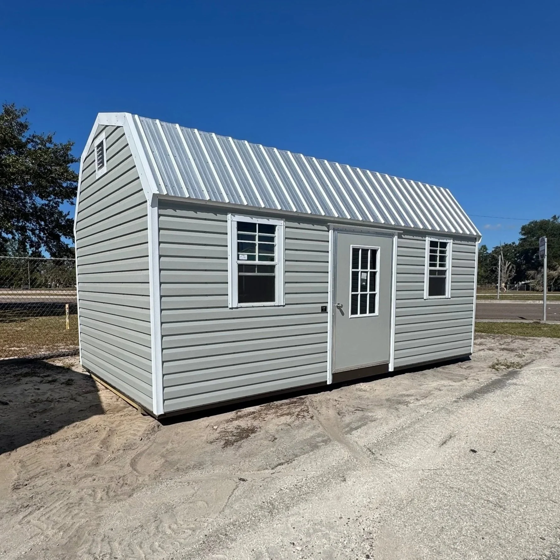 A small gray metal shed with white trim, two windows, and a door, situated on a gravel surface under a blue sky.