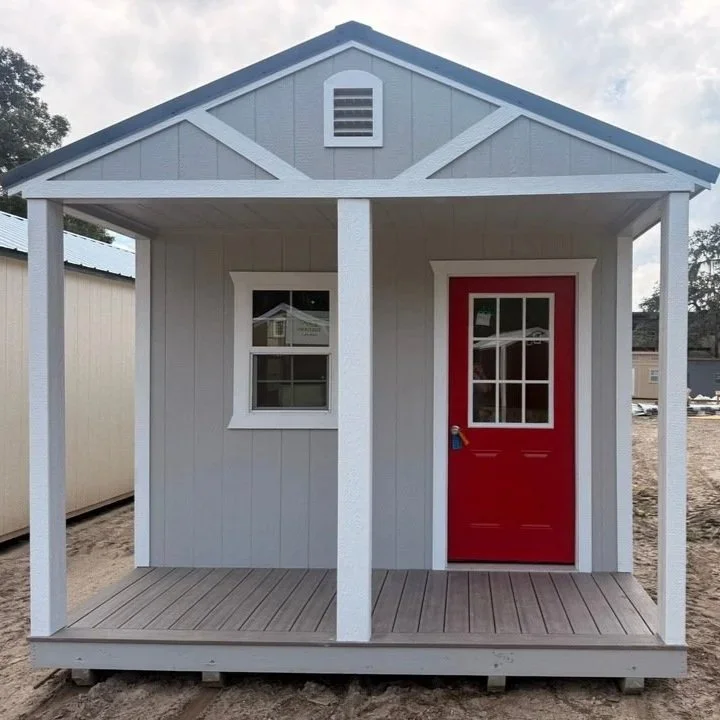 Small gray playhouse with a red door and a small window, built on a wooden platform with a porch, outdoors.