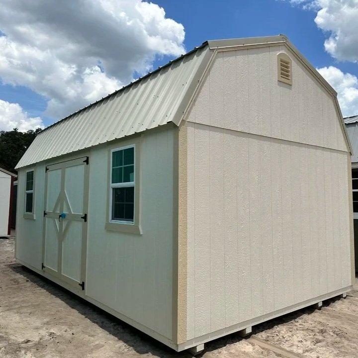 A small white storage shed with a metal roof, two windows, and a barn-style door, situated outdoors on a patch of dirt under a partly cloudy sky.