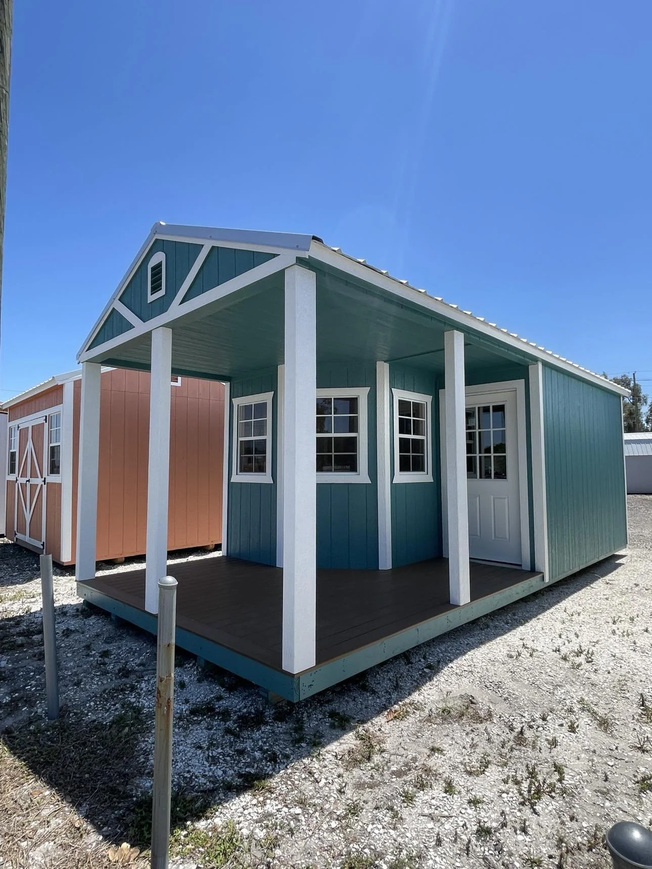 Colorful 12x24 cabin shed shed with blue walls, white trim, and a small porch, located outdoors on a gravel surface under a clear blue sky.