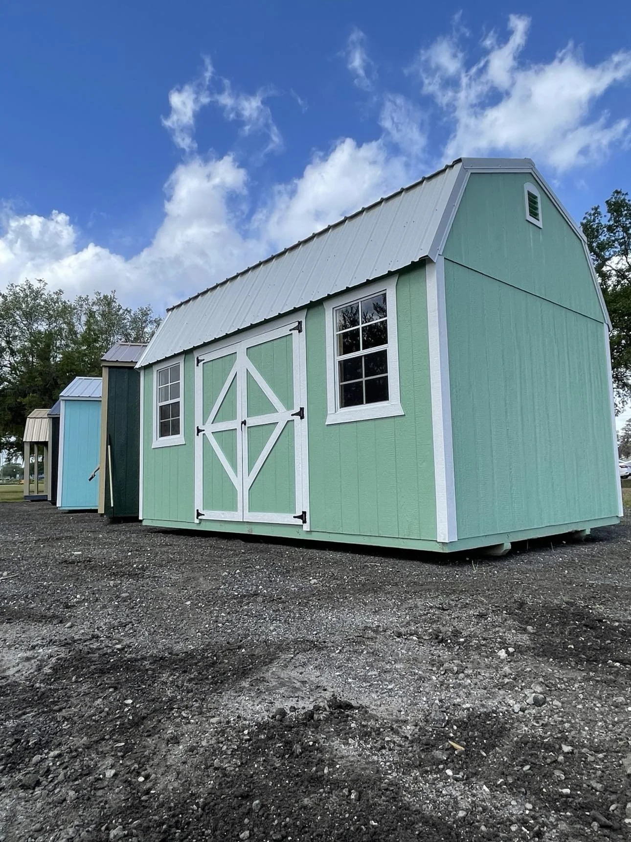 Colorful storage sheds on a gravel lot under a partly cloudy sky, featuring a green shed with white trim and black hardware, and smaller blue and dark green sheds in the background.