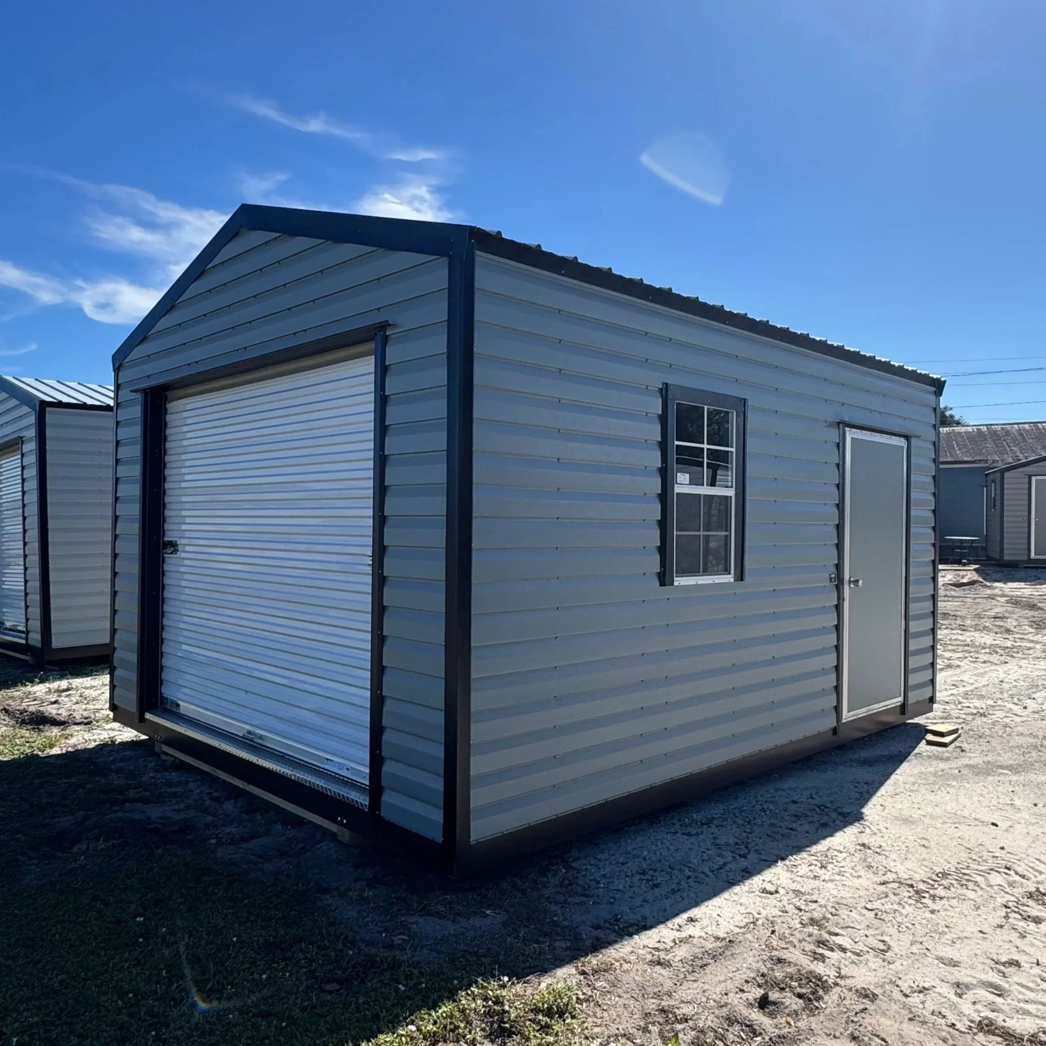 A small metal storage shed with a roll-up garage door, a side door, and a window, located outdoors on a dirt ground under a clear blue sky.