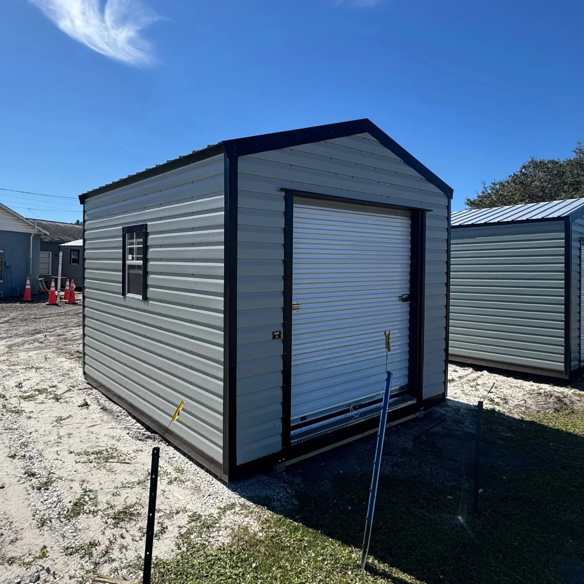 A small metal storage shed with a roll-up door and a black frame, set on a gravel surface under a clear blue sky.