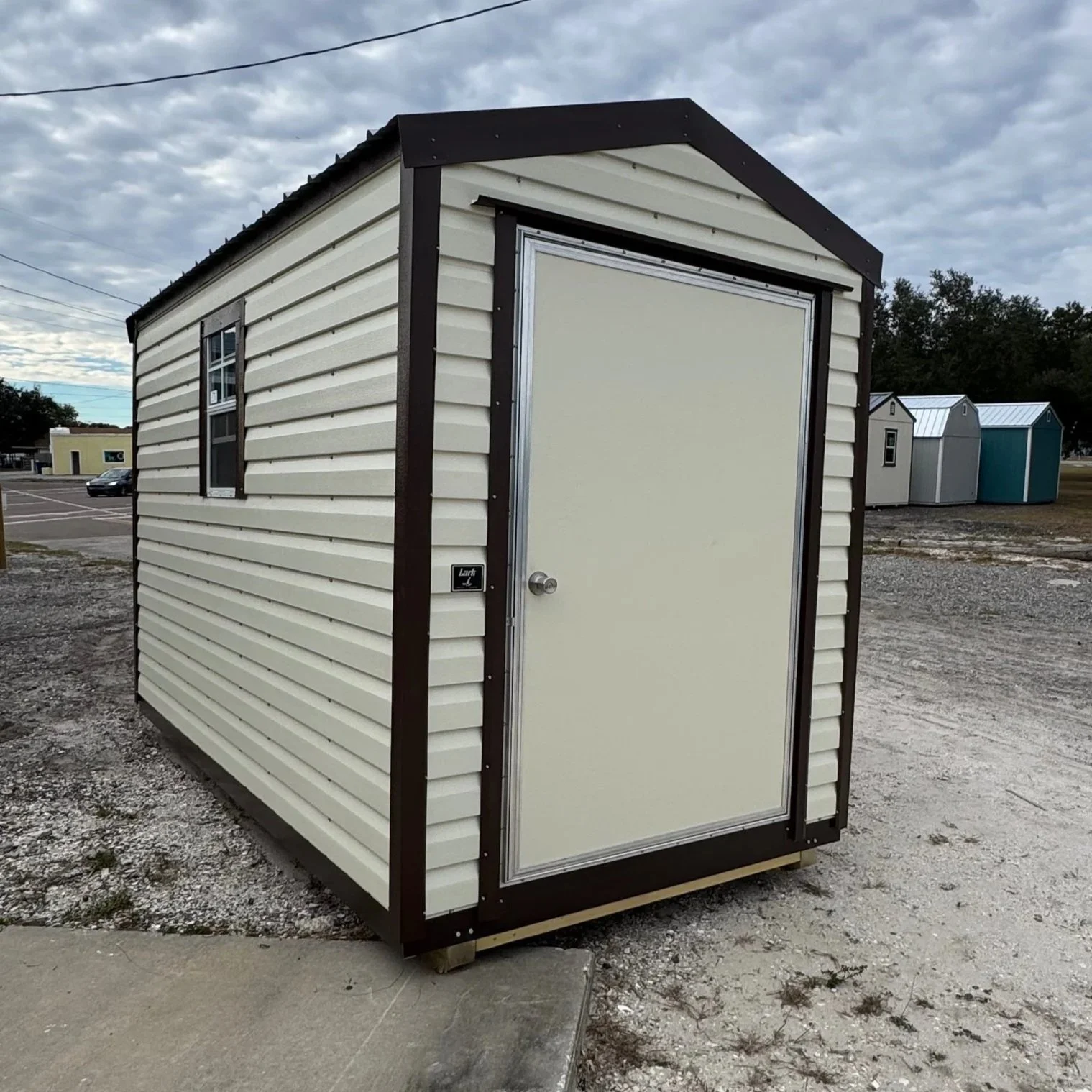 Small portable building with white siding, dark trim, and a door, situated outdoors on gravel with other similar structures in the background under a cloudy sky.