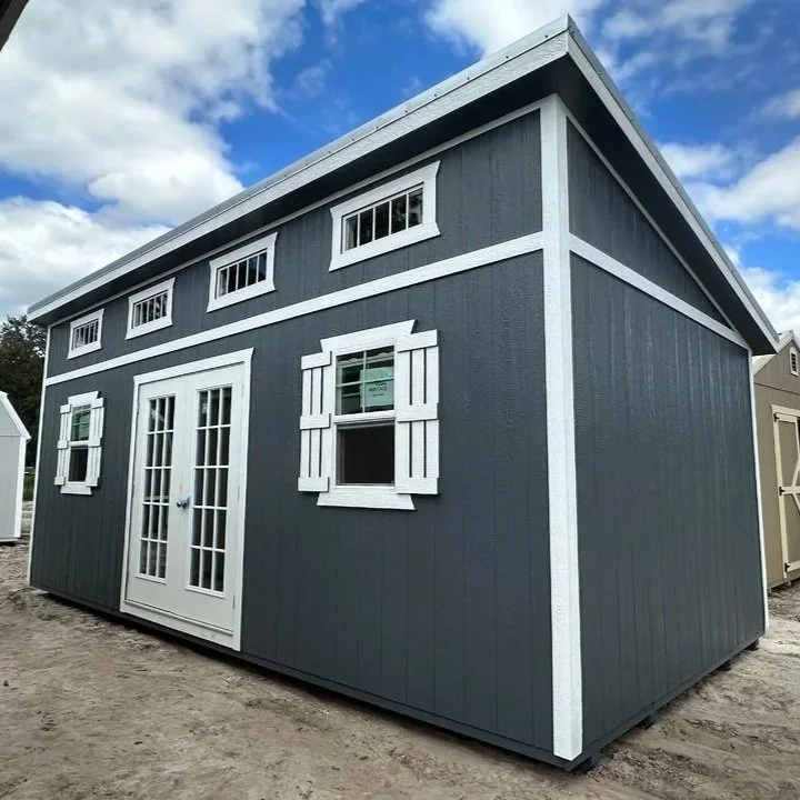 A small, dark blue shed with white trim, including a set of double glass doors, several windows with shutters, and a sloped roof, situated outdoors under a partly cloudy sky.