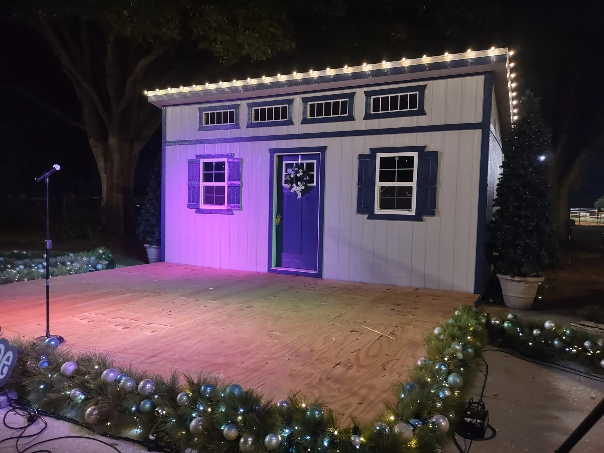 A small stage set up outdoors at night featuring a 12x20 shed with white and blue paneling, decorated with string lights along the roofline, and surrounded by Christmas garlands with silver and blue ornaments, with a microphone stand nearby.