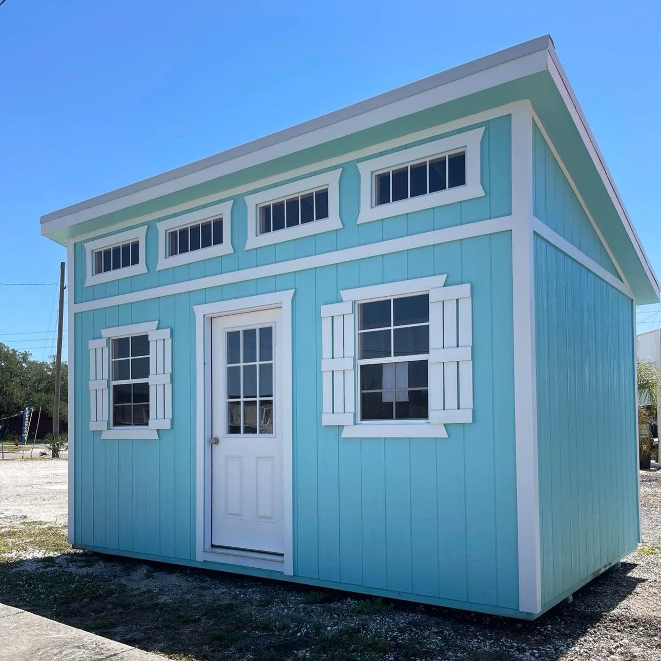 10x16 blue shed with white trim, two windows with white shutters, and a white door, set against a clear blue sky.