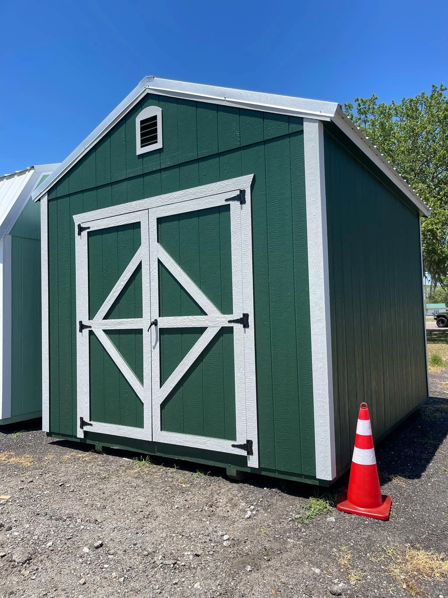 10x12 Green storage shed with white trim and double doors, concrete surface, orange traffic cone nearby, clear blue sky, trees in background.