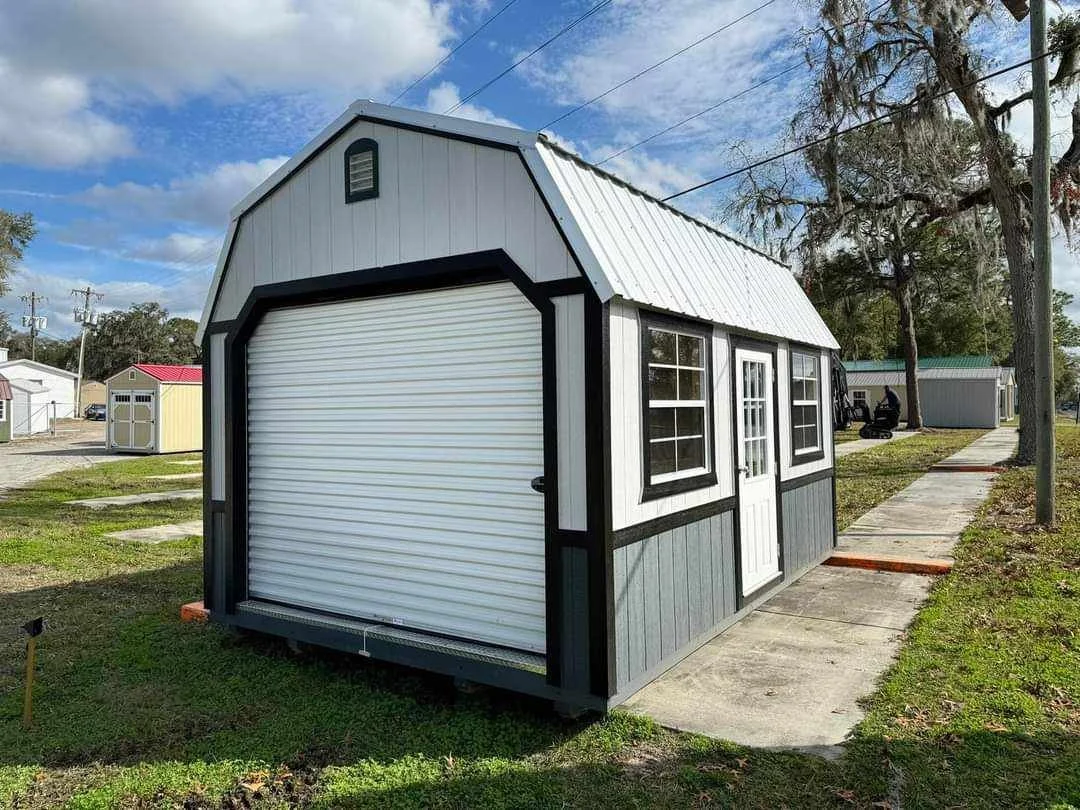 Small white and gray barn shed with a roll-up door, windows, and a door, situated on a concrete path in a yard.