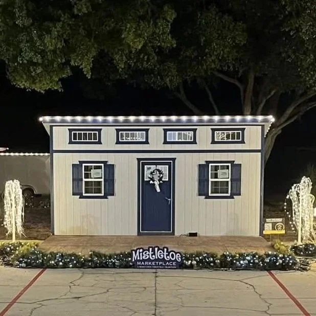 Small decorated house with string lights on the roof, white exterior with black accents, and a wreath on the door, set in a parking lot at night with mistletoe marketplace sign in front and two illuminated decorative spray arrangements.
