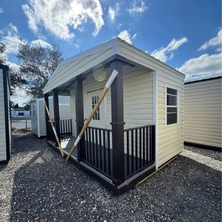 Small prefabricated house or tiny home under construction with support beams and a small front porch with black railing, set on a gravel lot under a partly cloudy sky.
