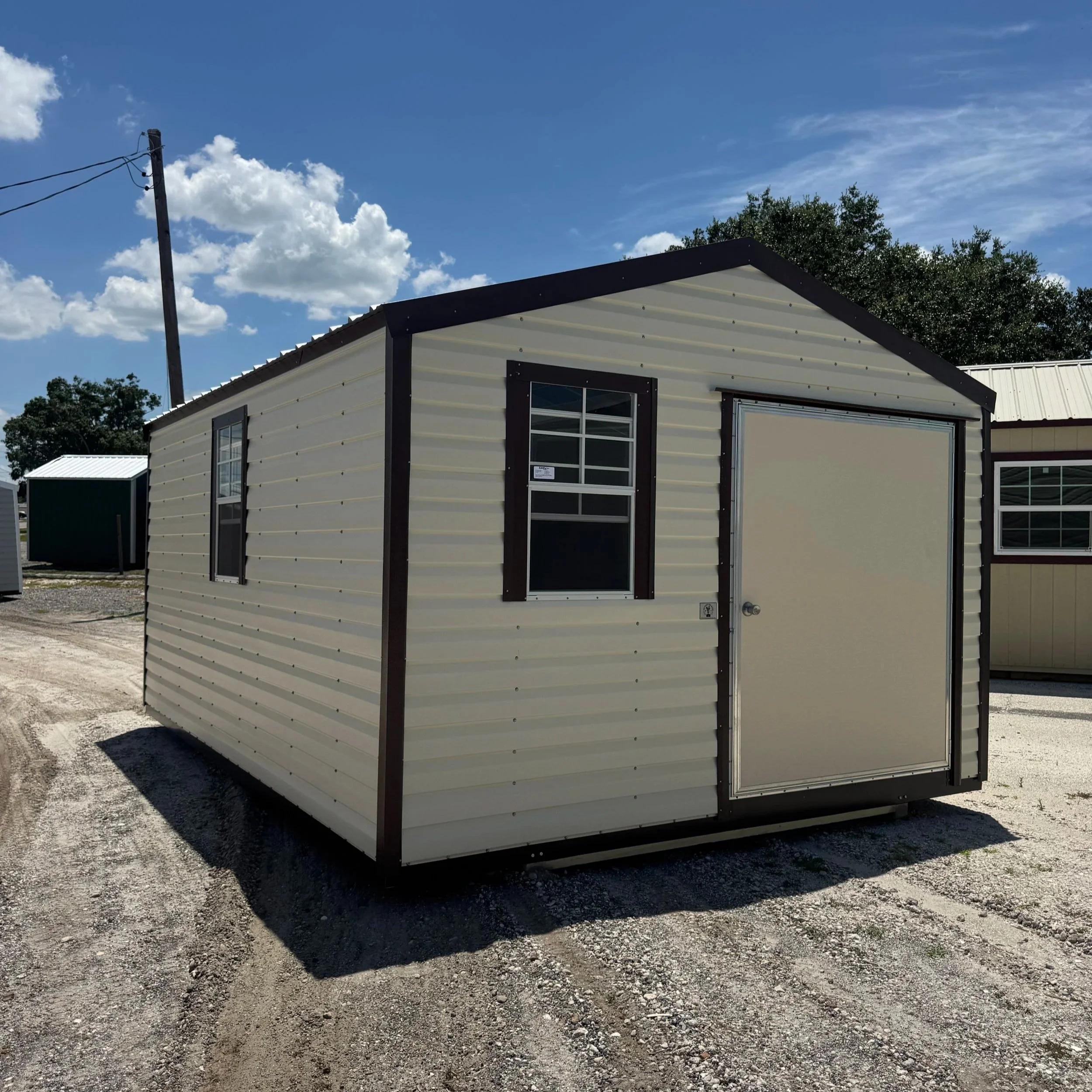 A small beige metal shed with dark trim, featuring a large door and two windows, situated on a gravel lot under a partly cloudy sky.