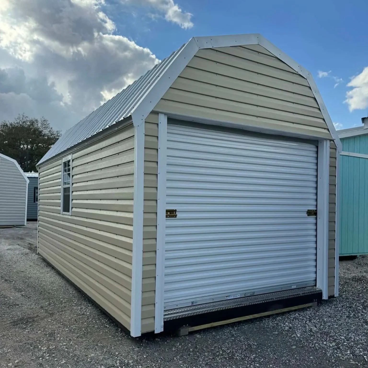 A beige metal storage shed with a white roll-up door, situated on a gravel lot under a partly cloudy sky.