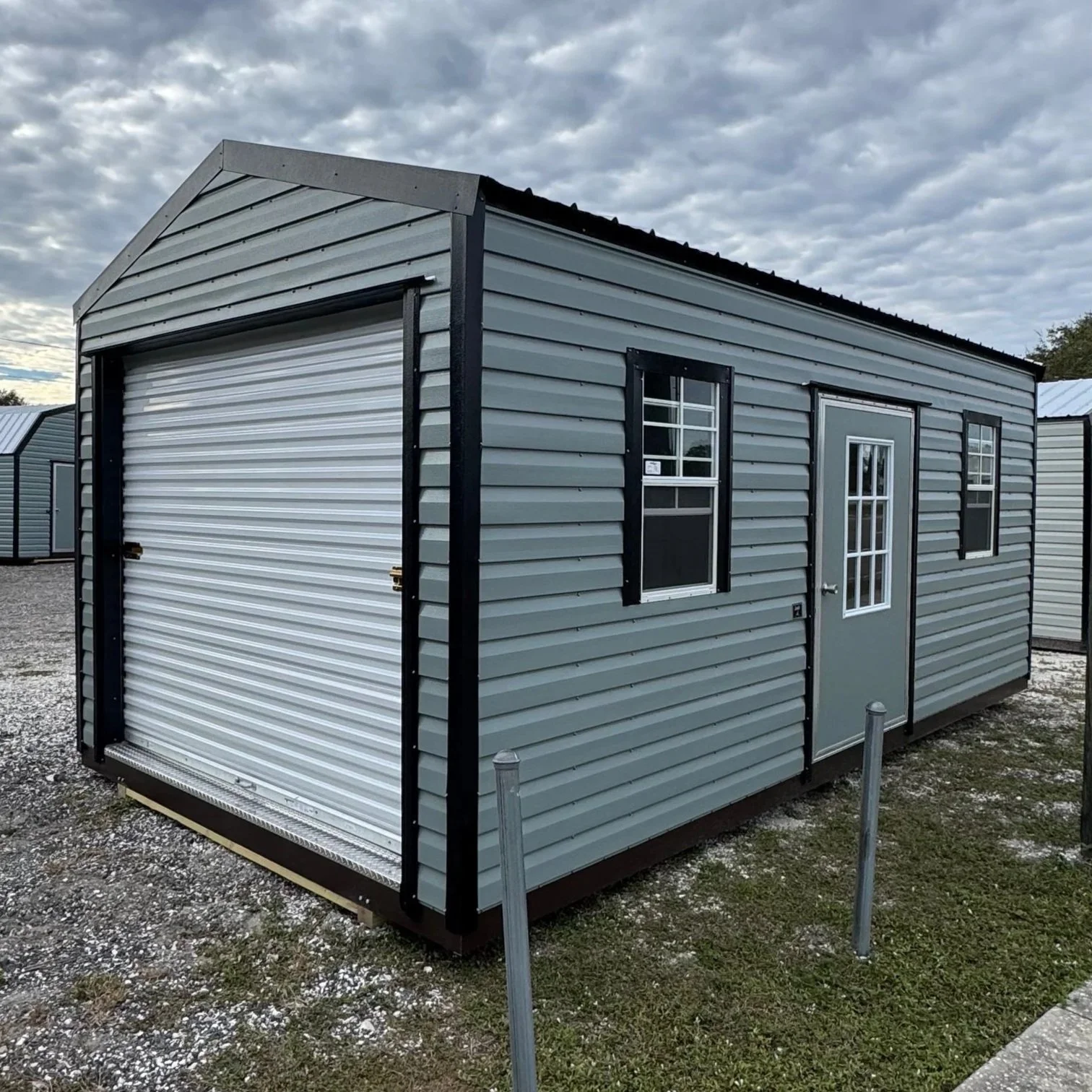 A gray metal storage shed with black trim, featuring a roll-up garage door, two small windows with black shutters, and a door with a windowpane, situated outdoors on a gravel surface with neighboring sheds and an overcast sky.