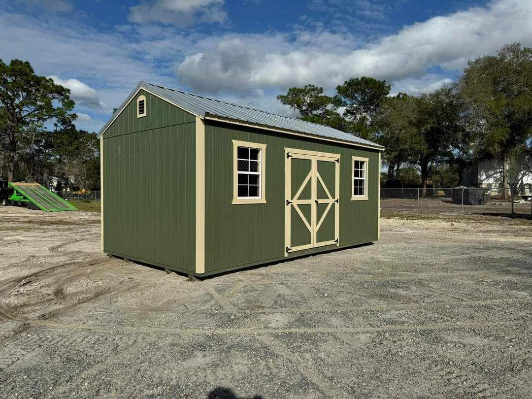 Green storage shed with beige trim and double doors on a gravel lot, with trees and a cloudy sky in the background.