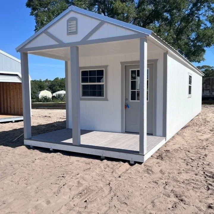 Small white cottage with a porch, double-hung windows, a door with a small window, and a sloped roof, set on a sandy lot.