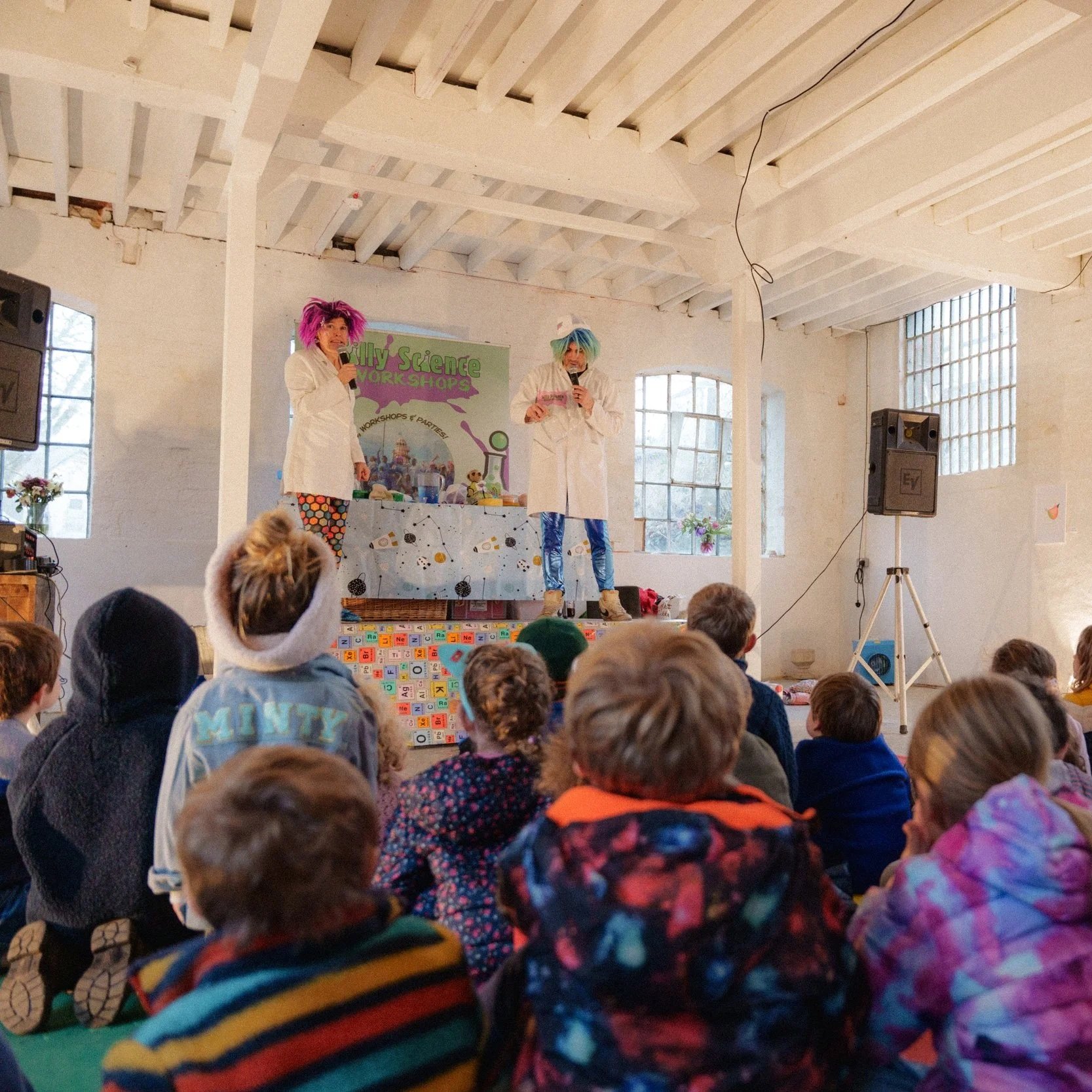 Two performers with colorful wigs and white lab coats presenting science shows to a large group of children in an industrial-style room with exposed beams and large windows. The children are seated on the floor, watching the presentation.