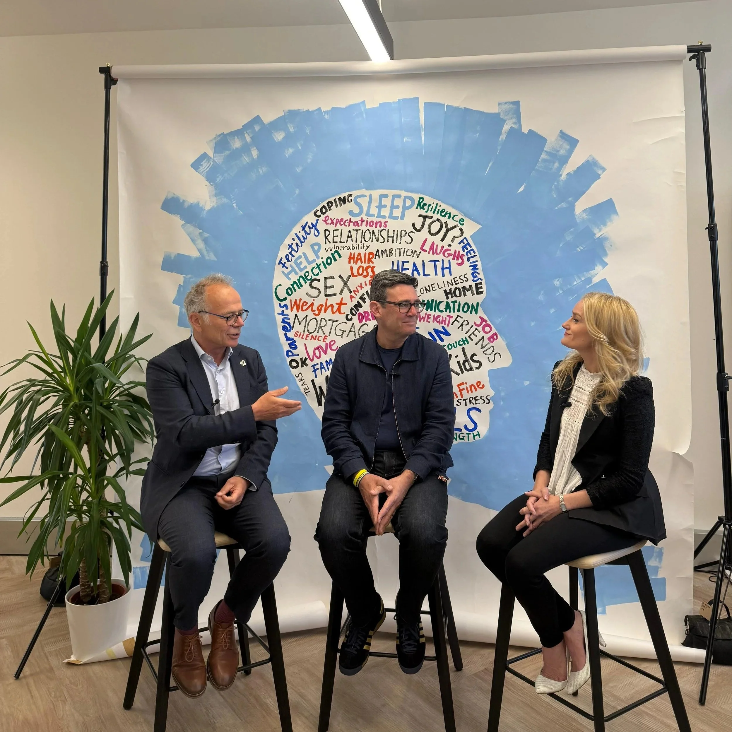 Group of four people sitting on stools in front of a backdrop covered in handwritten words related to health, relationships, and emotions, likely at an event or discussion.