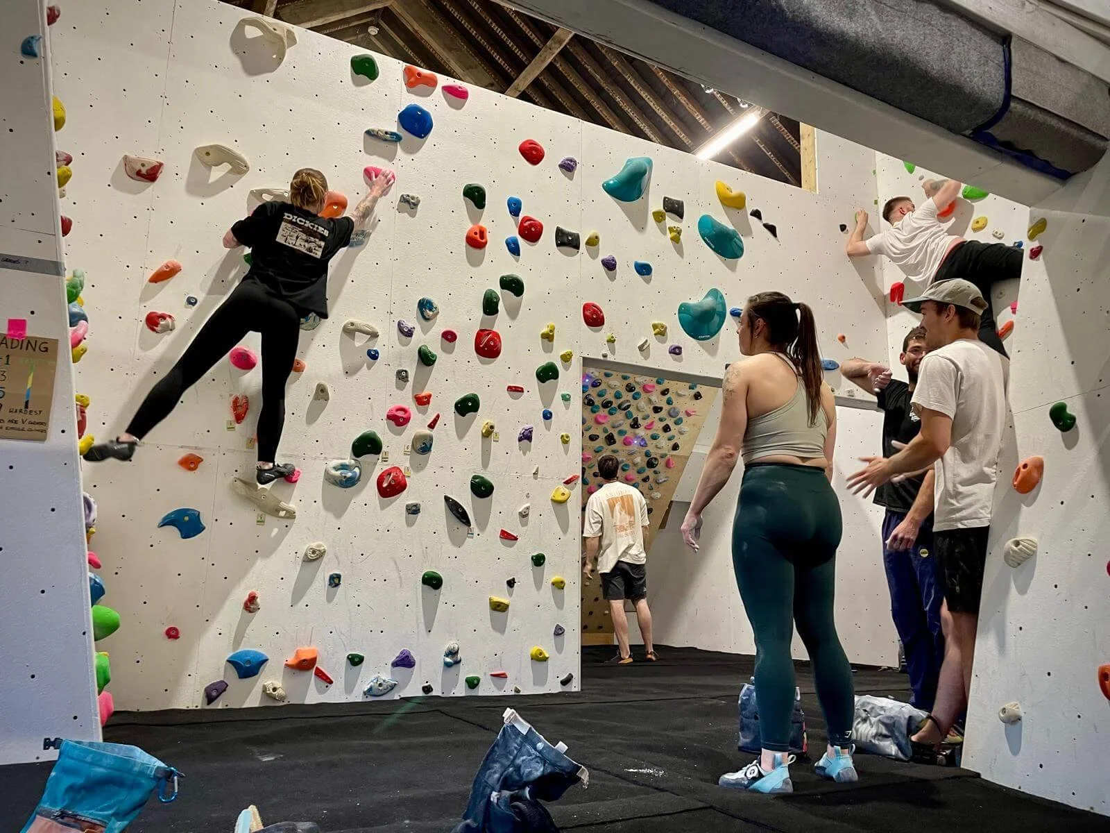 People indoor rock climbing at a climbing gym, with several individuals climbing on the wall and others observing or waiting their turn.