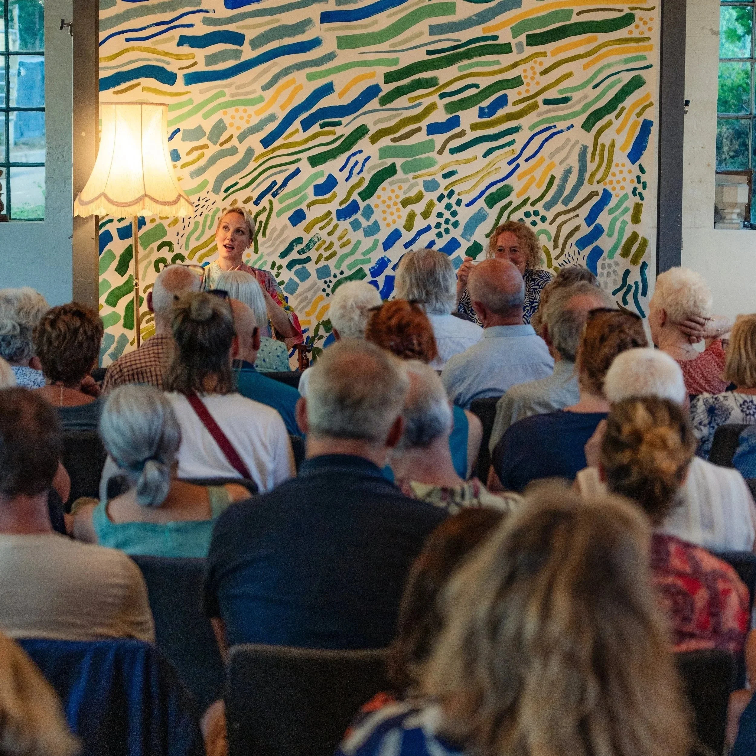 A group of people attending a presentation in a room with large windows and a colorful abstract mural wall. A woman is speaking, and there is a standing lamp next to her.