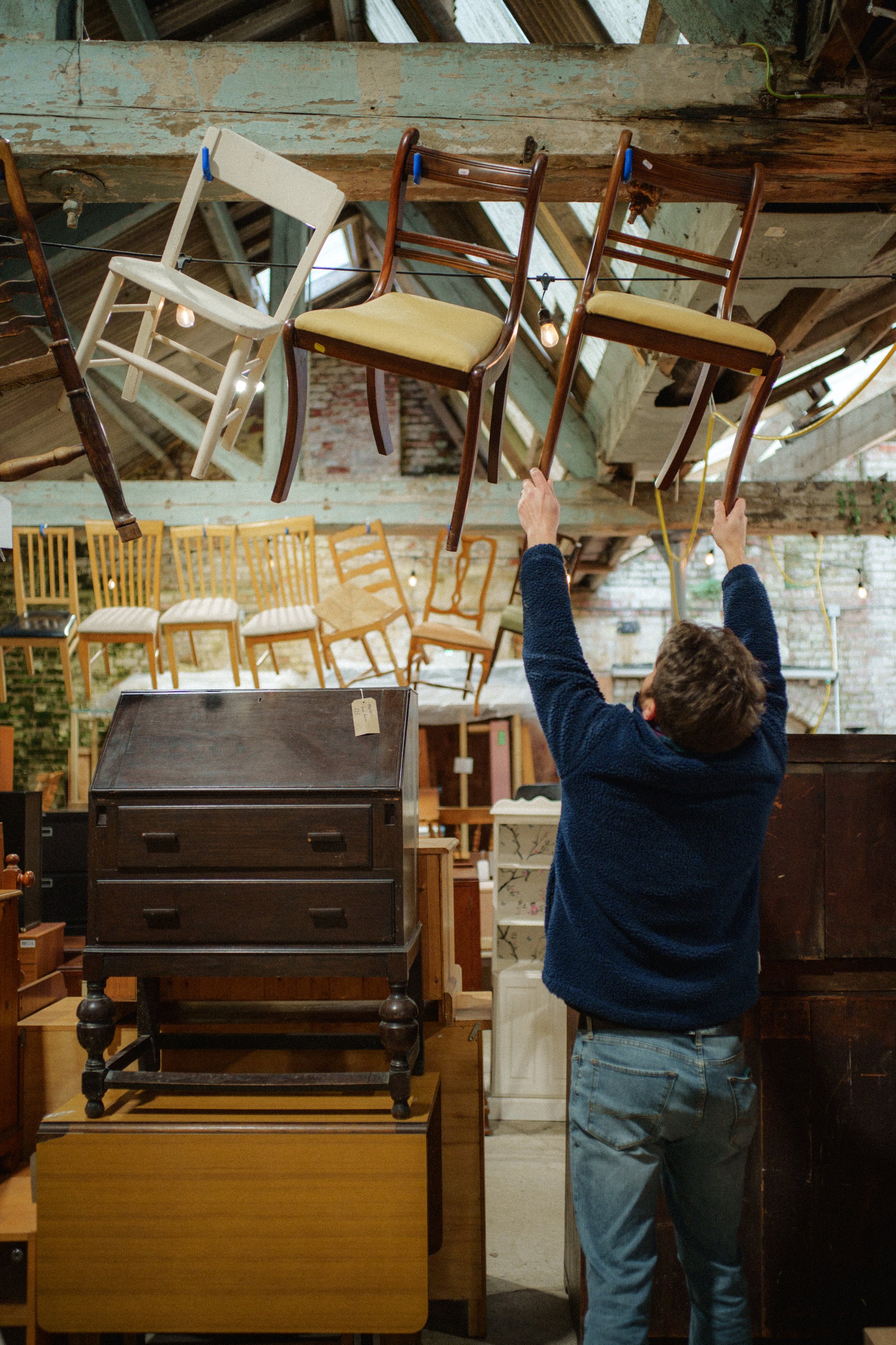 A person is hanging multiple chairs from a wire or string in a large vintage or antique furniture store with wooden beams and a brick wall. The chairs include a white wooden chair and two dark wood chairs with yellow cushions. The person's face is not visible, and they are wearing a dark blue sweater and jeans.