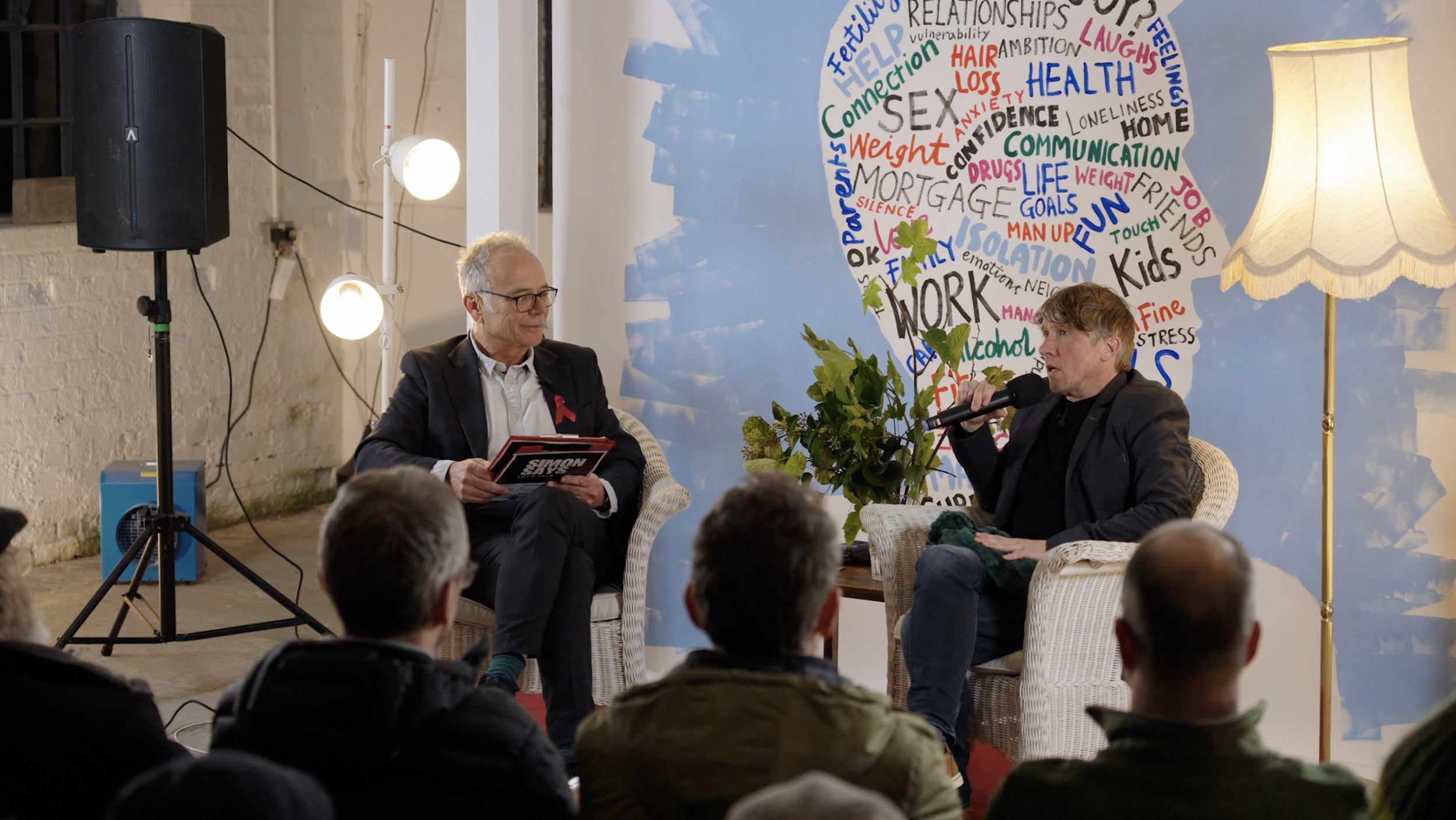A man with glasses and gray hair wearing a dark blazer and holding a book, seated next to a different man with brown hair speaking into a microphone, during a discussion or interview. They are in a room with a large colorful word cloud shaped like a head on the wall behind them, and a standing lamp providing light. Several people are seated in front of them, watching the conversation.