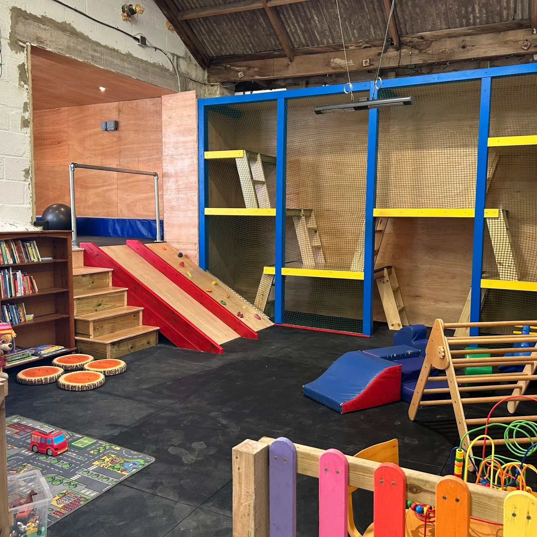 Indoor children's play area with climbing structures, a slide, soft flooring, bookshelves, and toys.