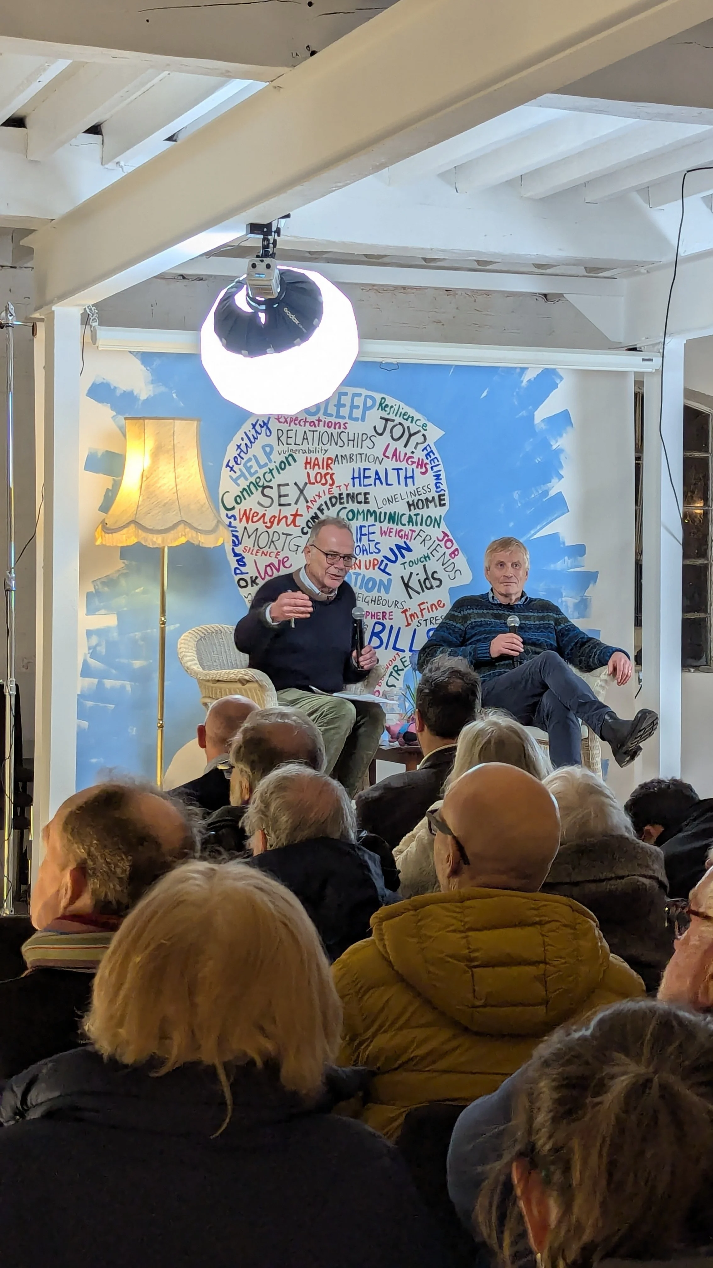 Two men are sitting on stage in front of an audience, participating in a discussion. One man is speaking into a microphone while the other is holding a microphone and listening. Behind them is a colorful wall with words related to relationships, health, and life experiences arranged within a lightbulb-shaped design. The setting appears to be indoors with stage lighting and an audience of various ages seated in front.