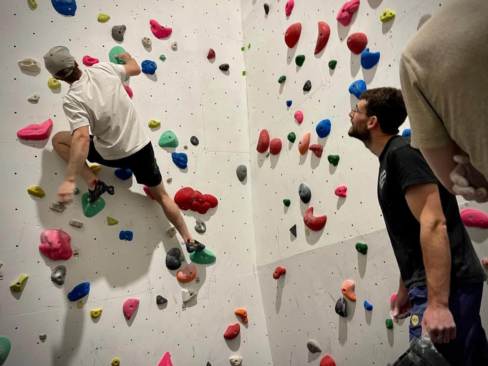 People at an indoor rock climbing gym. One climber, wearing a white shirt and black shorts, is climbing on a wall with colorful holds. A man with glasses, wearing a black shirt, is watching and smiling.